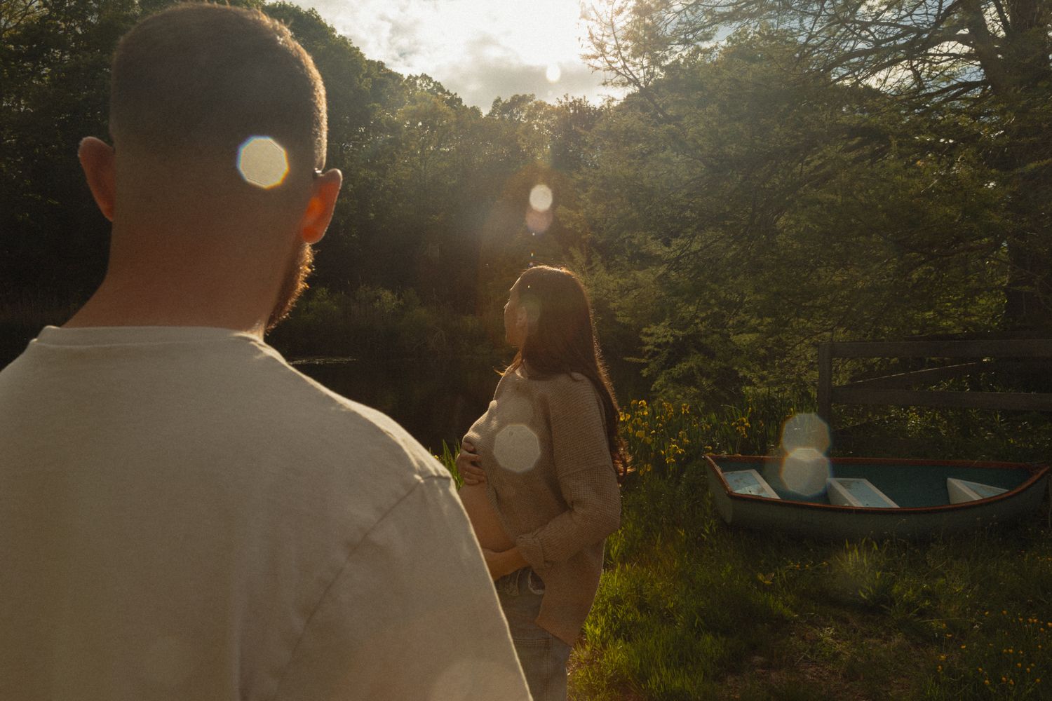Couple in sun-dappled forest clearing, pregnant woman holding belly near canoe at golden hour.