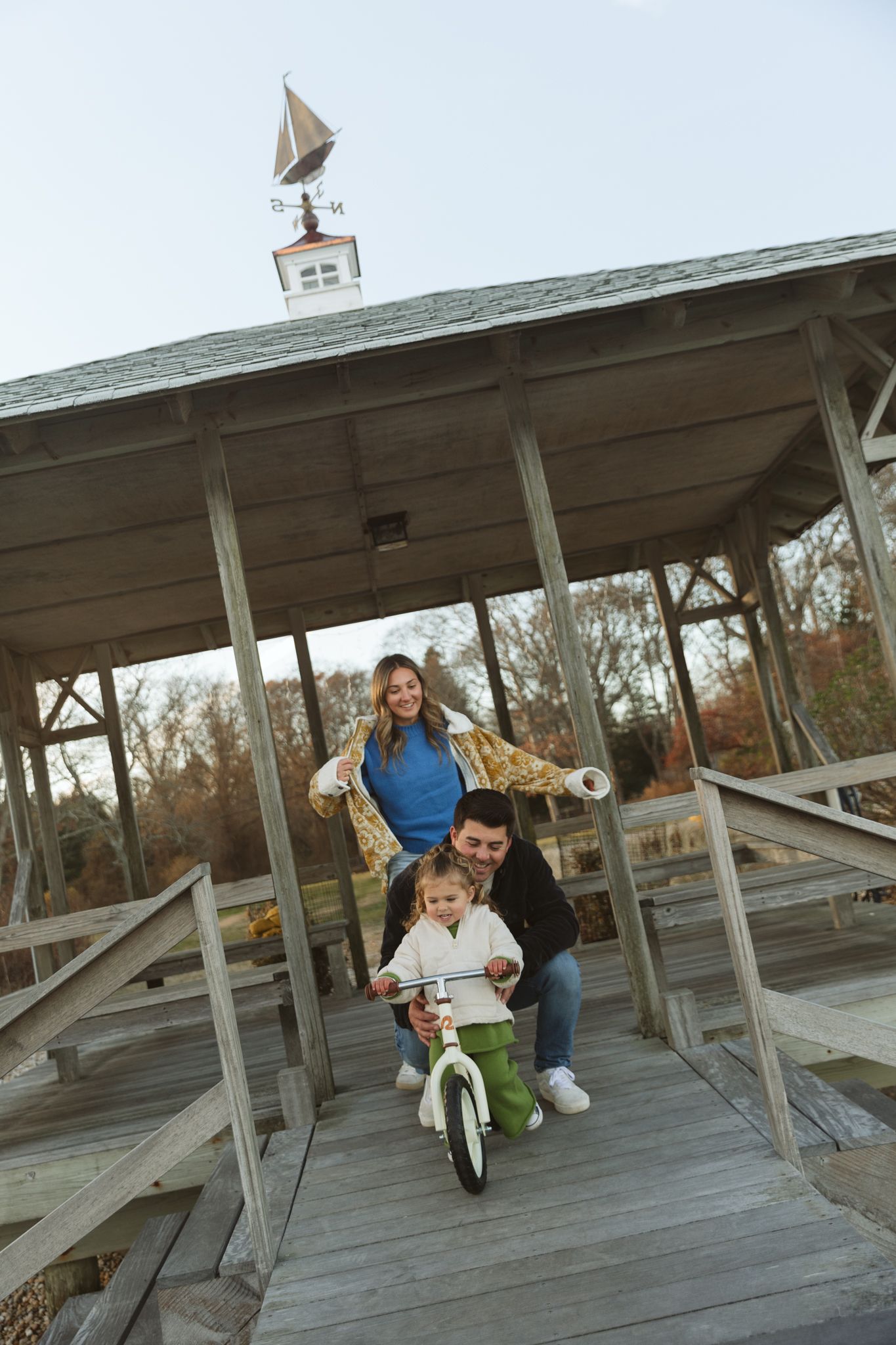 Family helping young child ride balance bike on wooden gazebo deck in autumn park.
