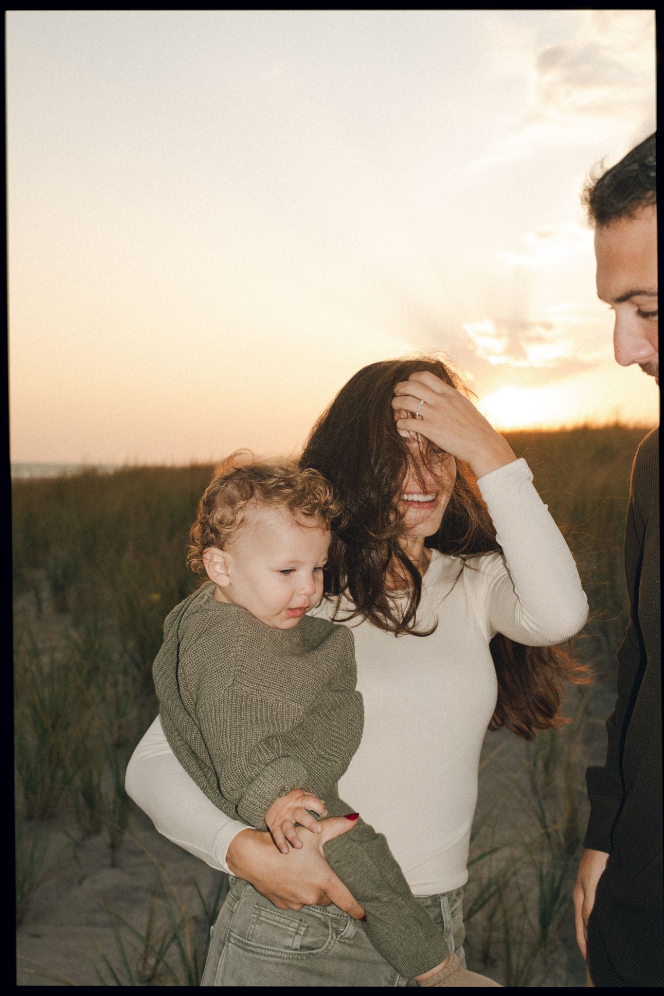 A smiling woman holds a toddler in a green sweater on a beach at sunset, with a man partially visible beside them.