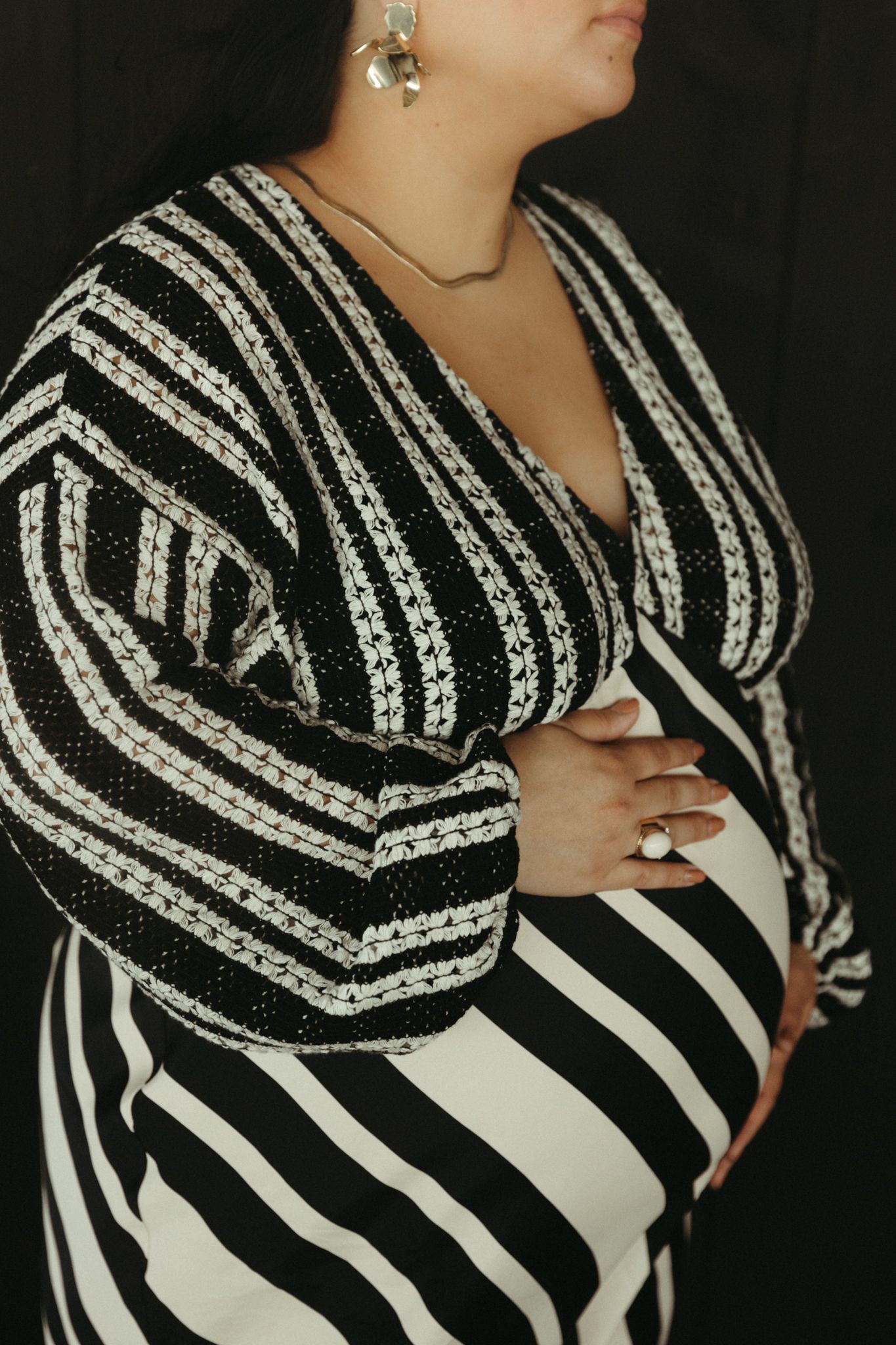 Pregnant woman in black and white striped dress cradling baby bump, wearing silver jewelry against dark background.