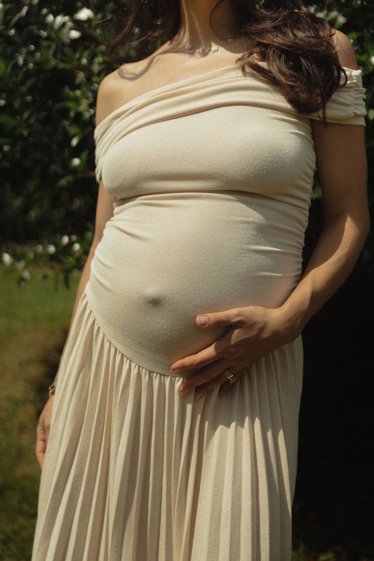 Pregnant woman in cream off-shoulder pleated dress cradling baby bump outdoors in garden setting.