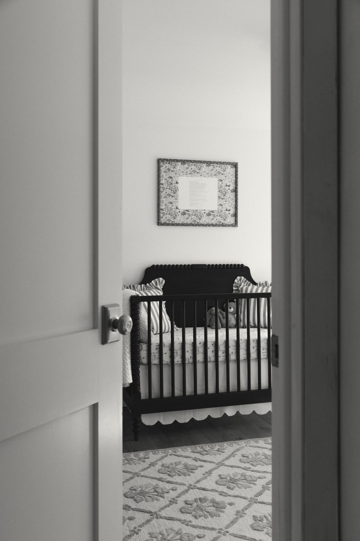 Black and white view of nursery through doorway showing crib and patterned rug.