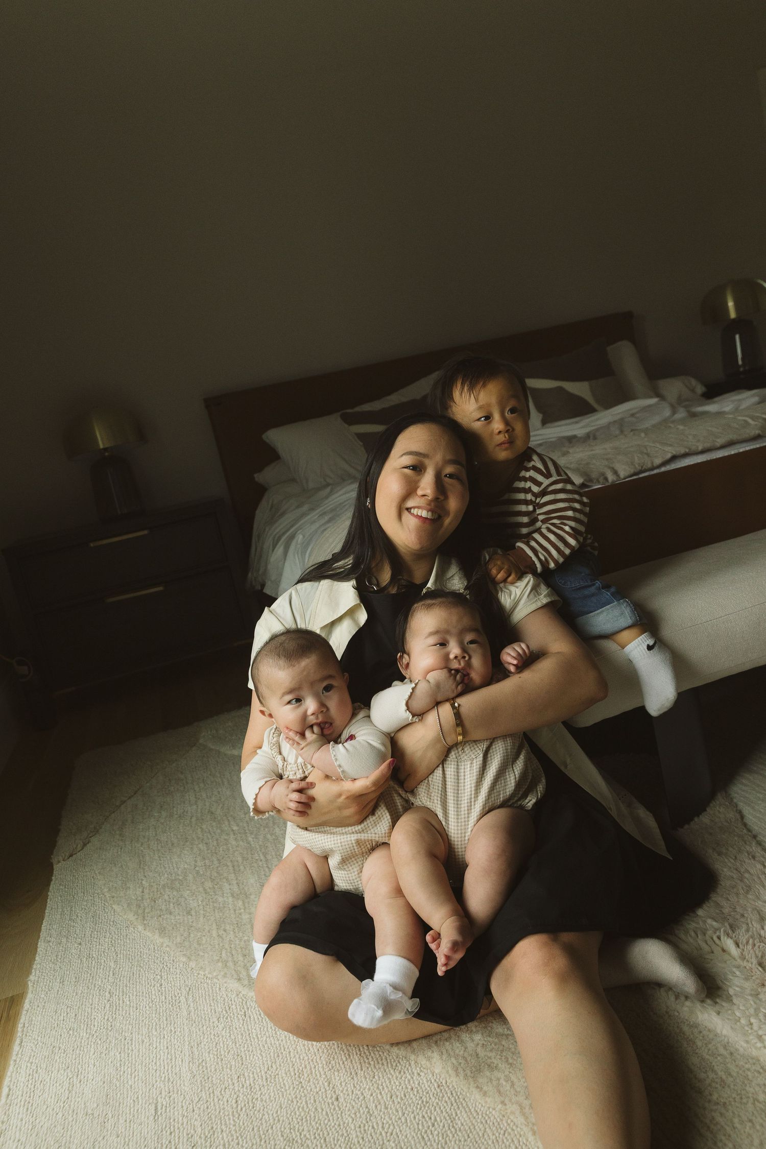 Smiling Asian mother sitting on bedroom floor holding three young babies and toddler in warm natural light.