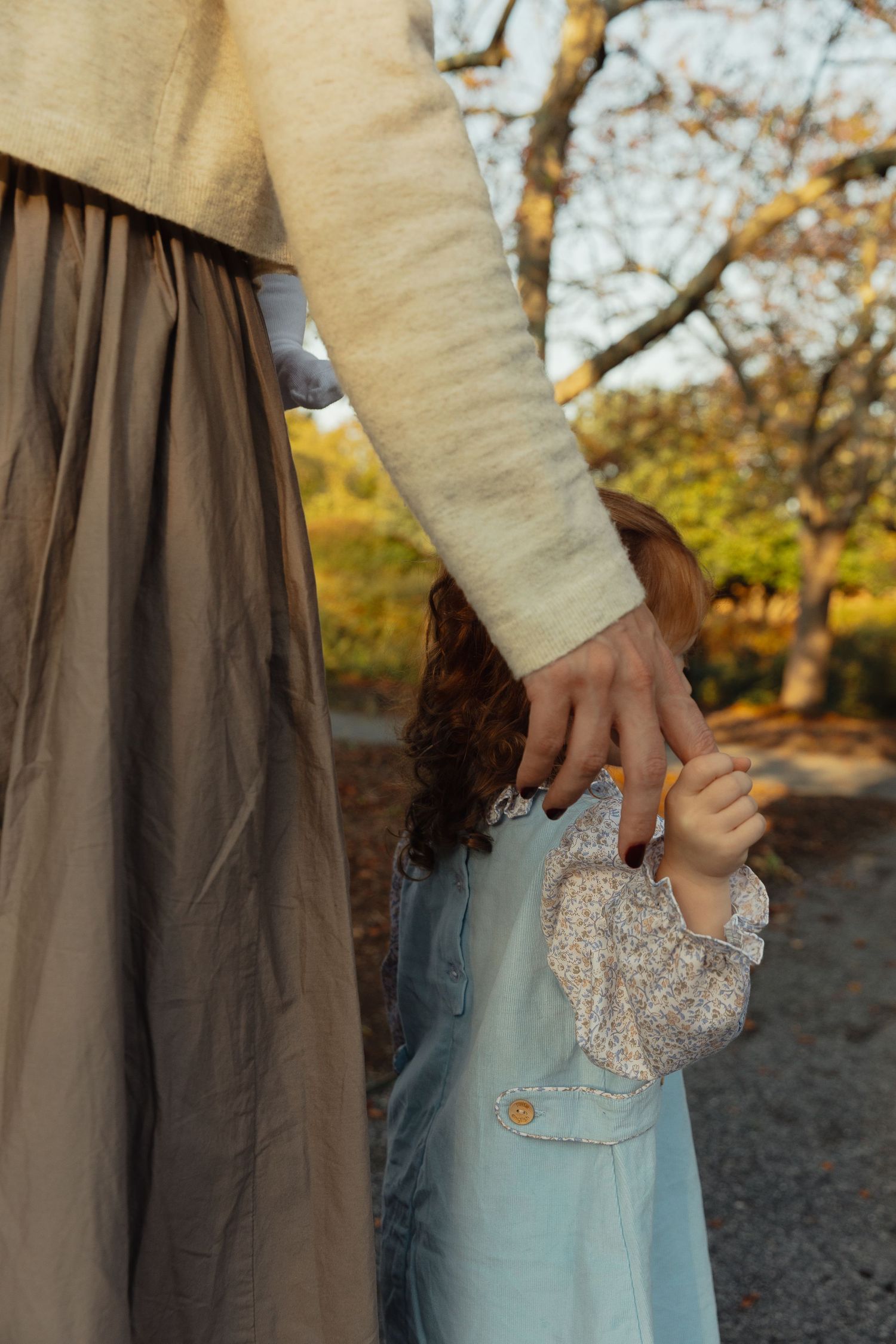 A mother holds her young child's hand while walking outdoors in autumn, both wearing soft neutral tones.