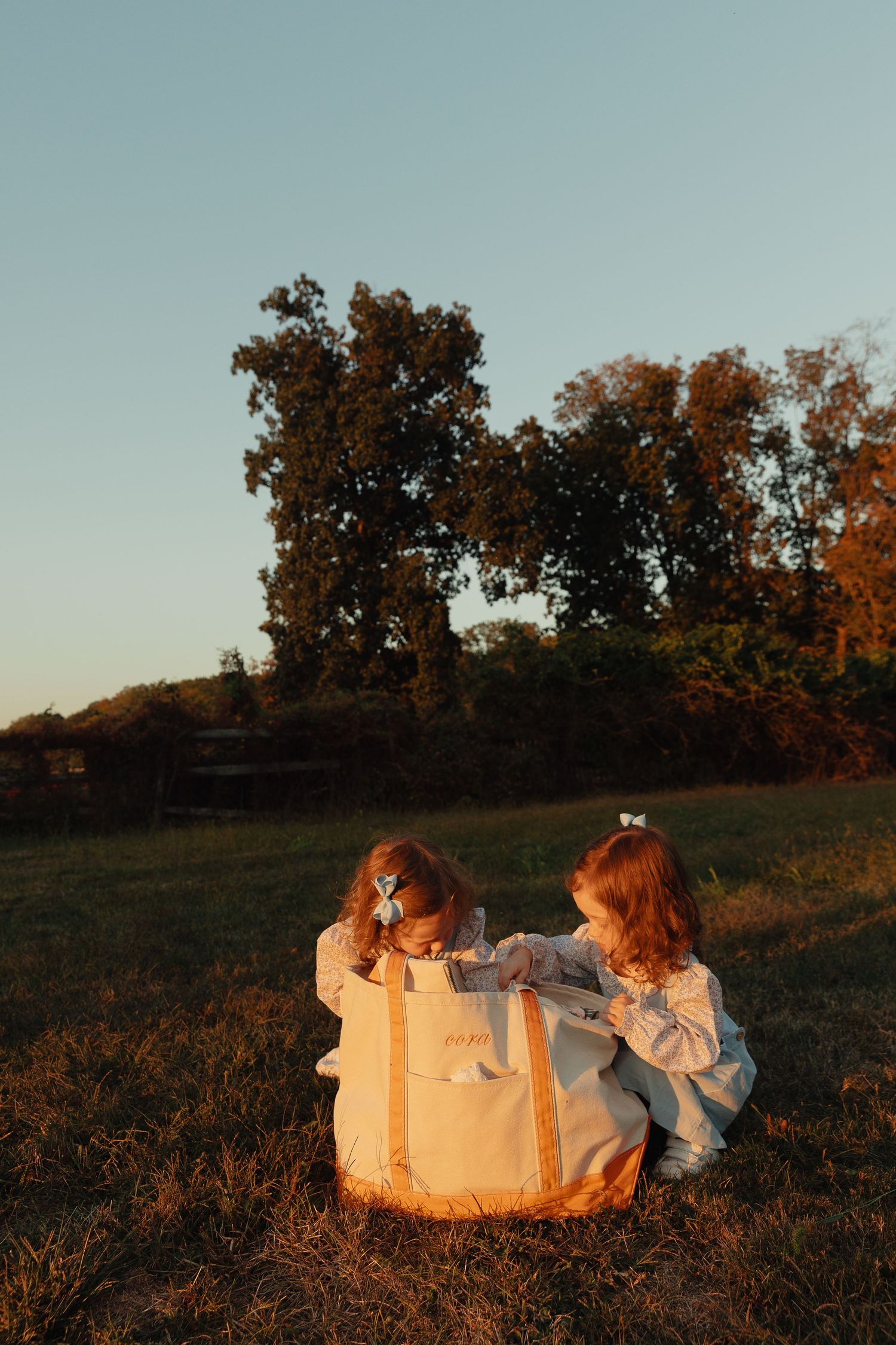 Two young girls in white dresses sit on grass at golden hour, looking into a large canvas tote bag outdoors.