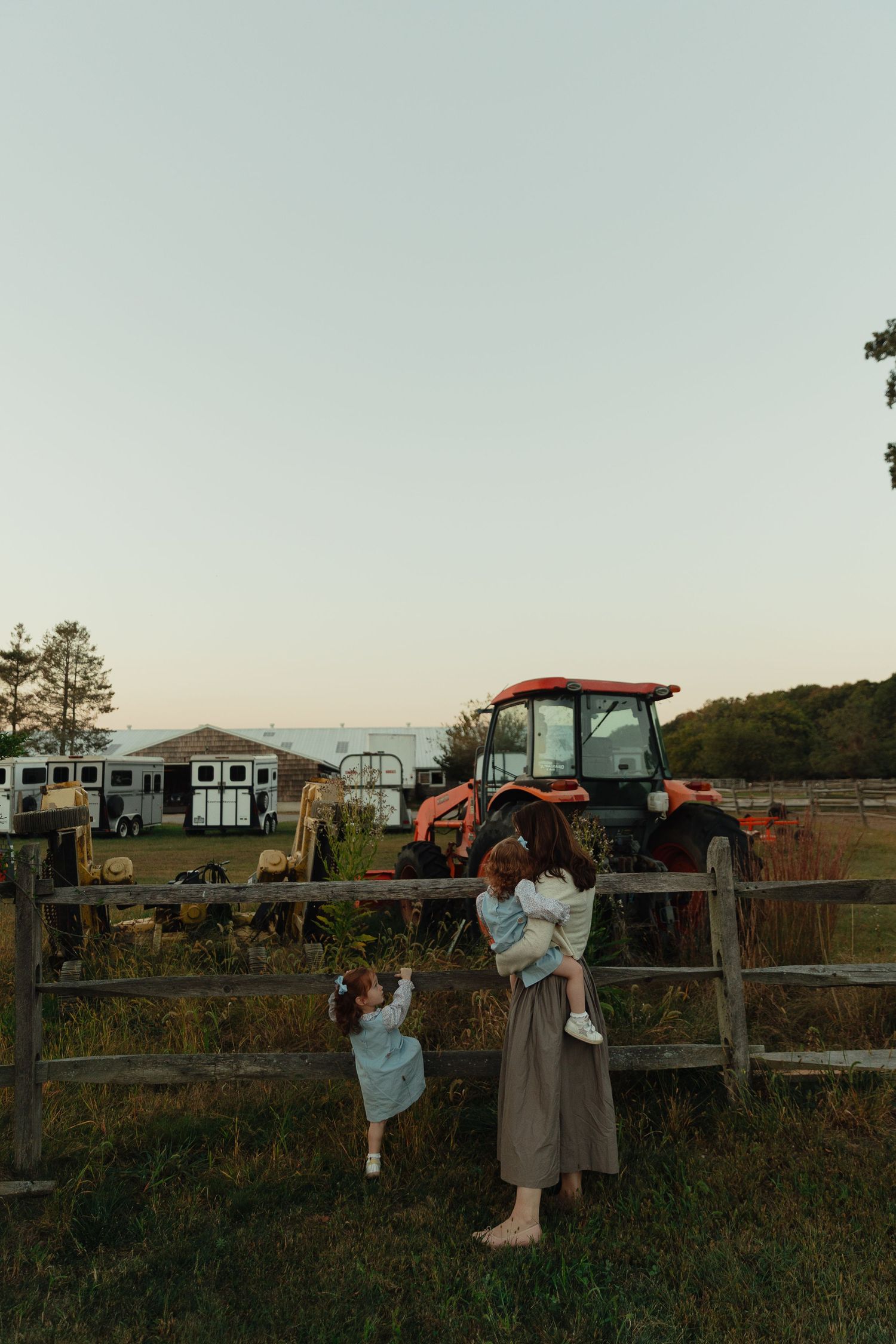 A mother holds her child by a wooden fence on a farm at dusk, with a red tractor and trailer visible behind them.