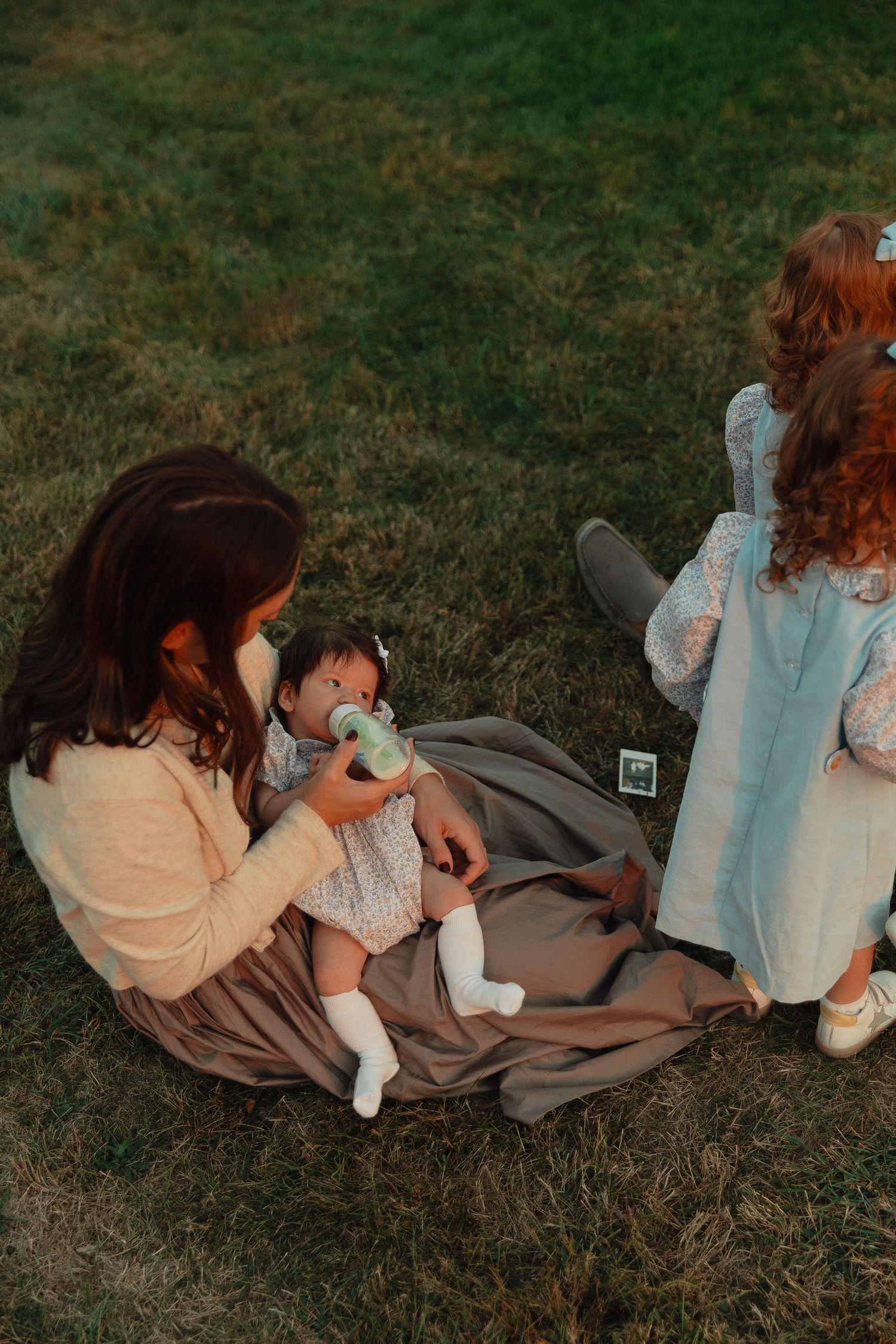A mother feeds her baby with a bottle while sitting on a blanket in the grass at dusk.