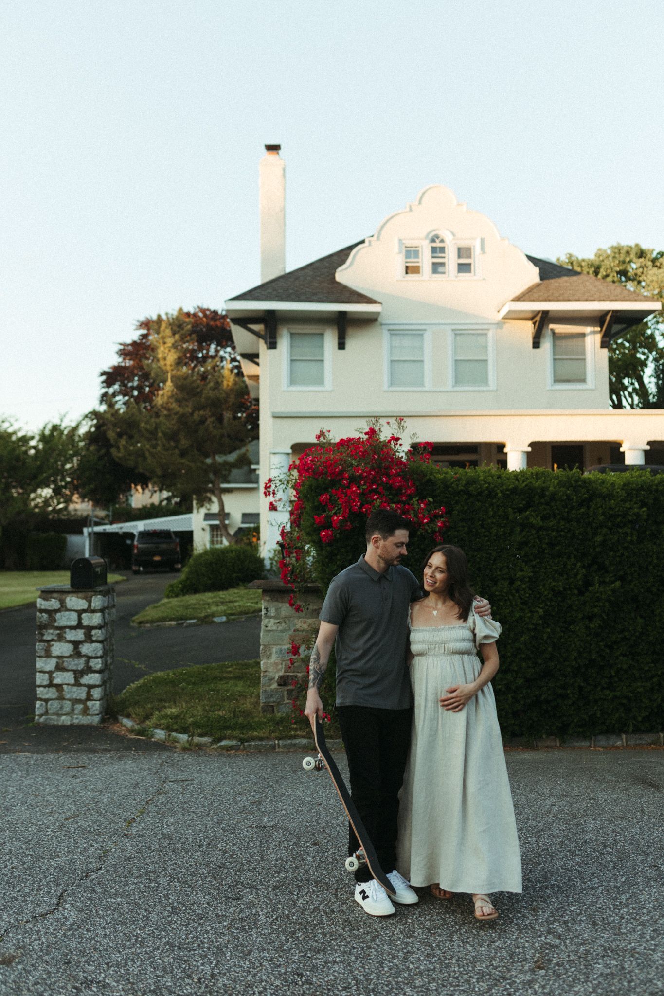 Pregnant couple standing in front of a white house, man holding a skateboard, woman in a light green maternity dress.