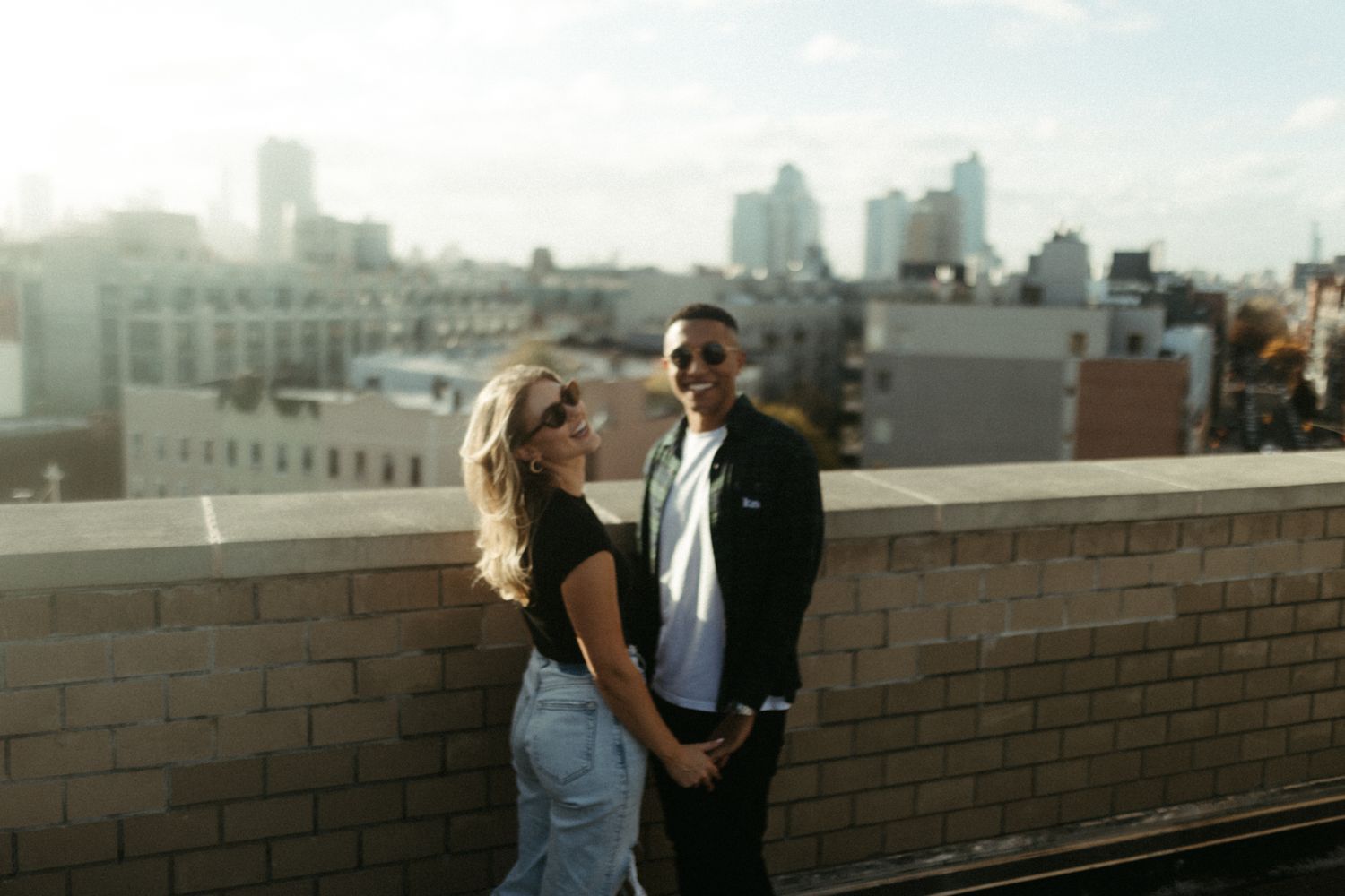 Couple holding hands on a rooftop with a city skyline backdrop on a sunny day.