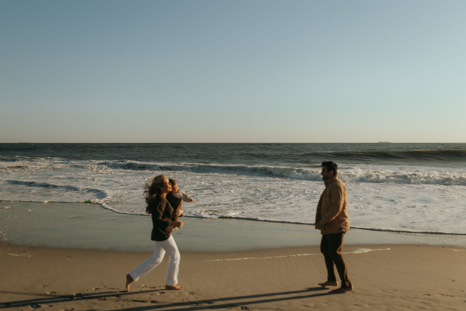 Two people walk along a beach with waves crashing in the background at sunset.