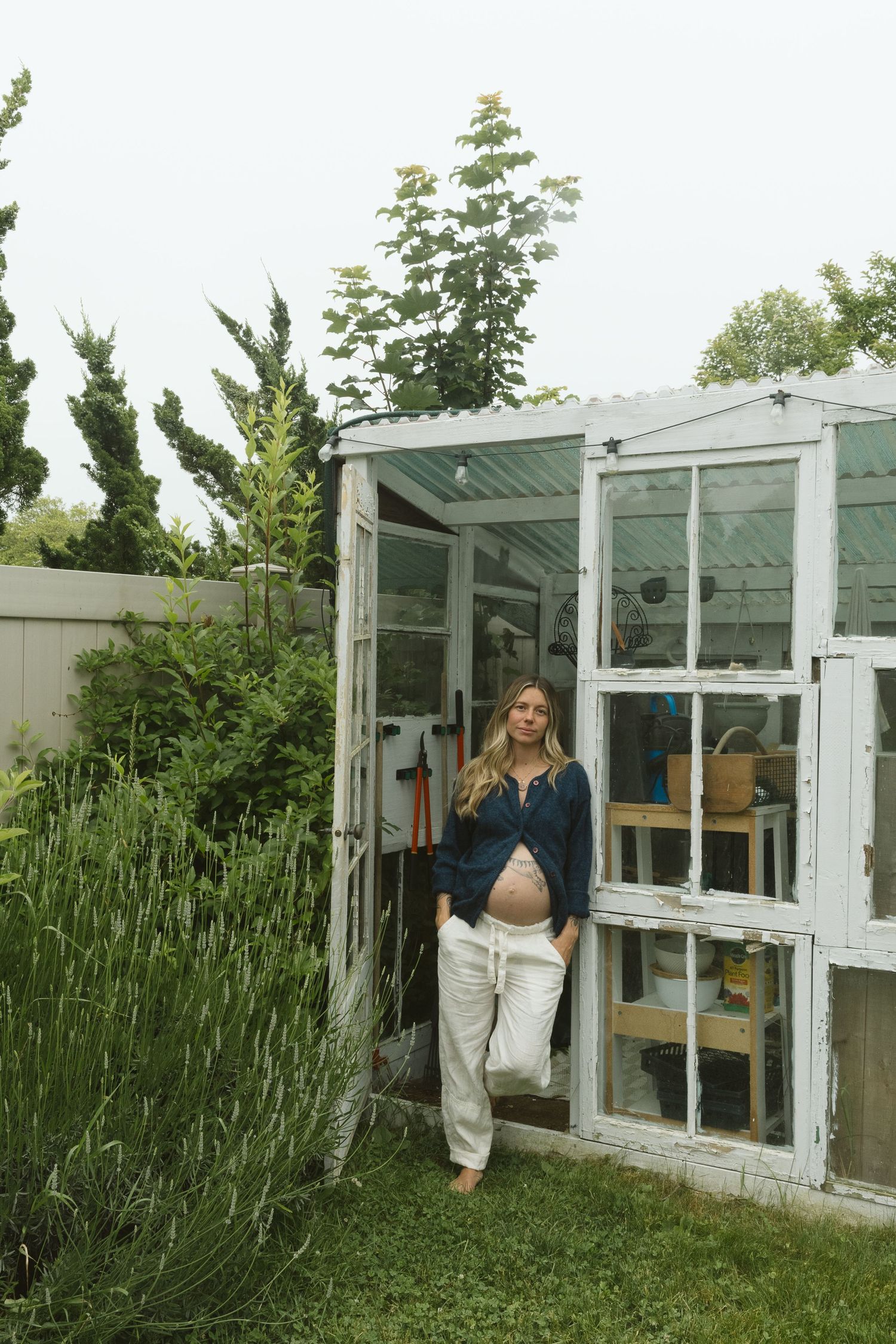 Expectant mother in white pants and navy top standing in greenhouse doorway.