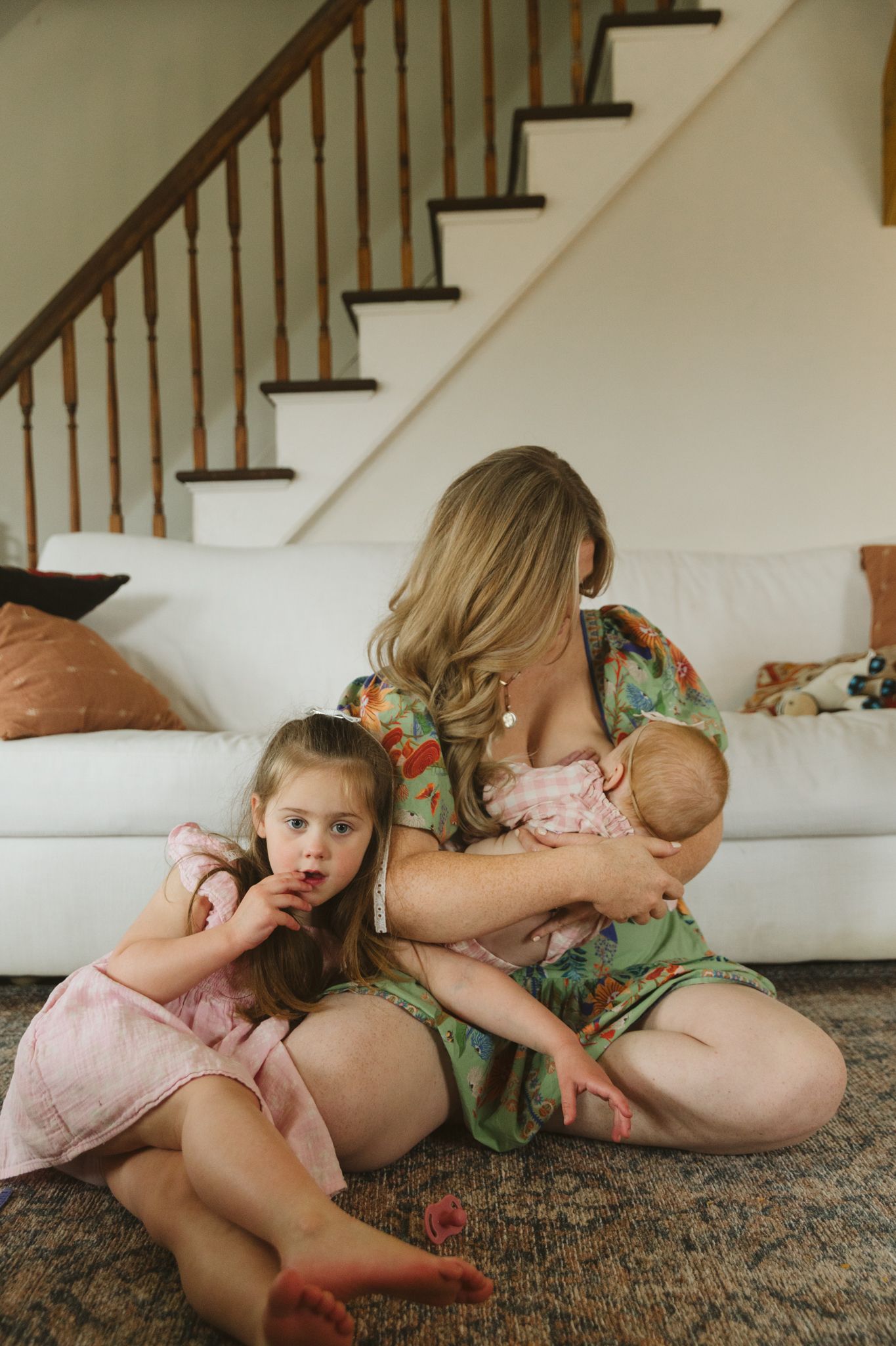Older sisters sitting while mom breastfeeds baby sister in an in-home East Quogue family session, capturing love and family connection
