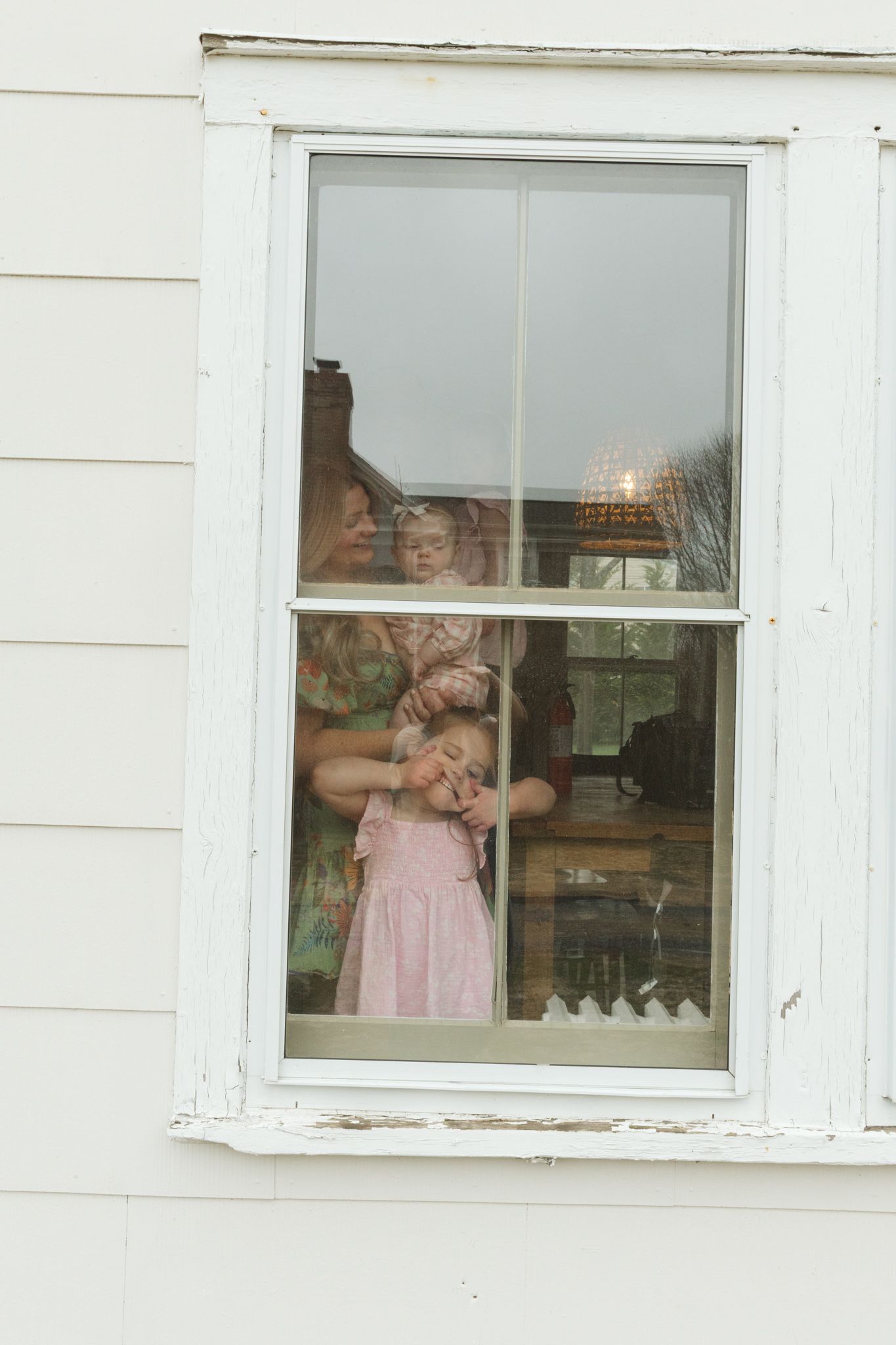 Young girl in pink dress standing at vintage white window with adult visible behind her.