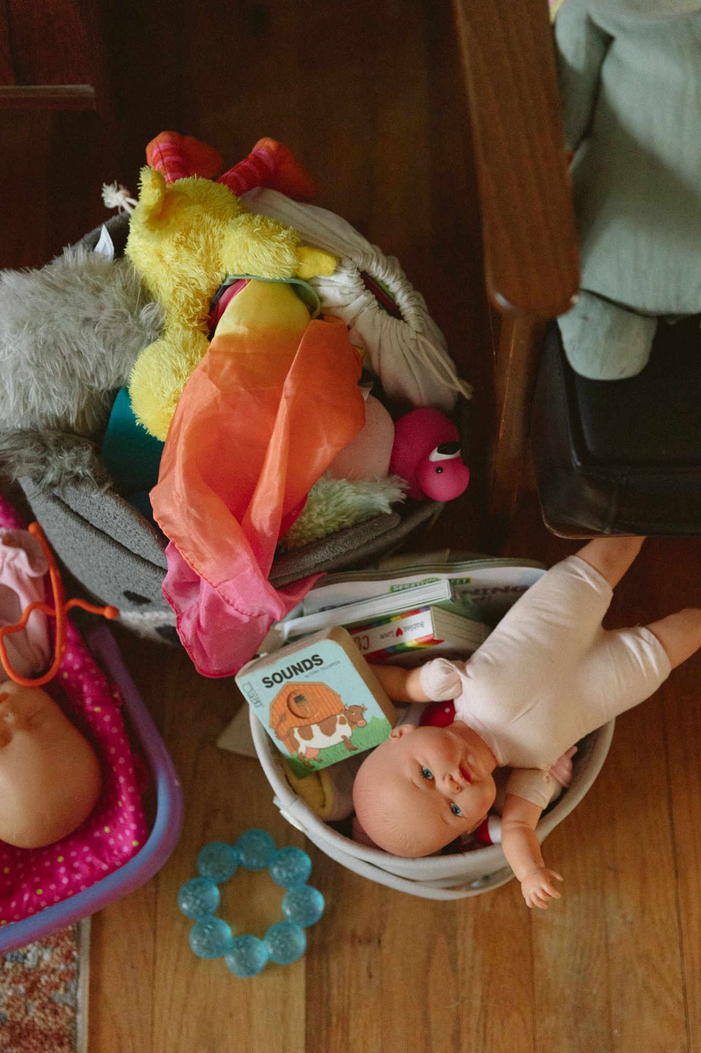 Overhead view of children's toys including stuffed animals, baby doll, board book, and teething ring on wooden floor.