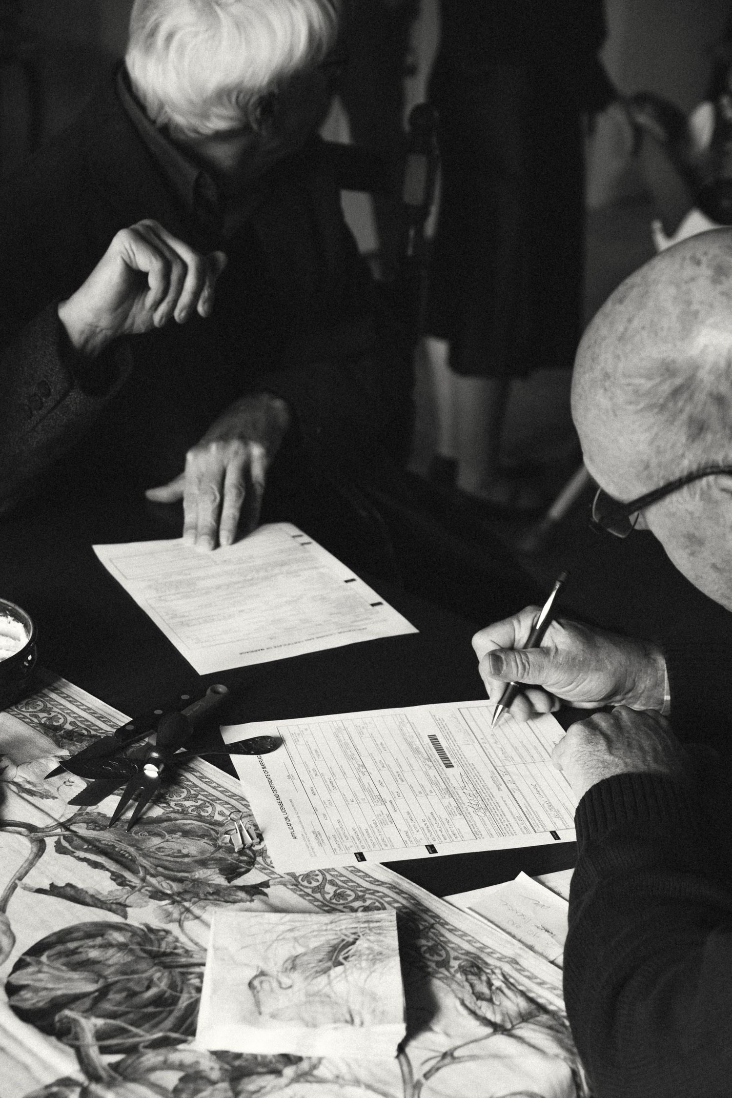 Black and white photo of elderly people signing documents at a table covered with papers and artwork.