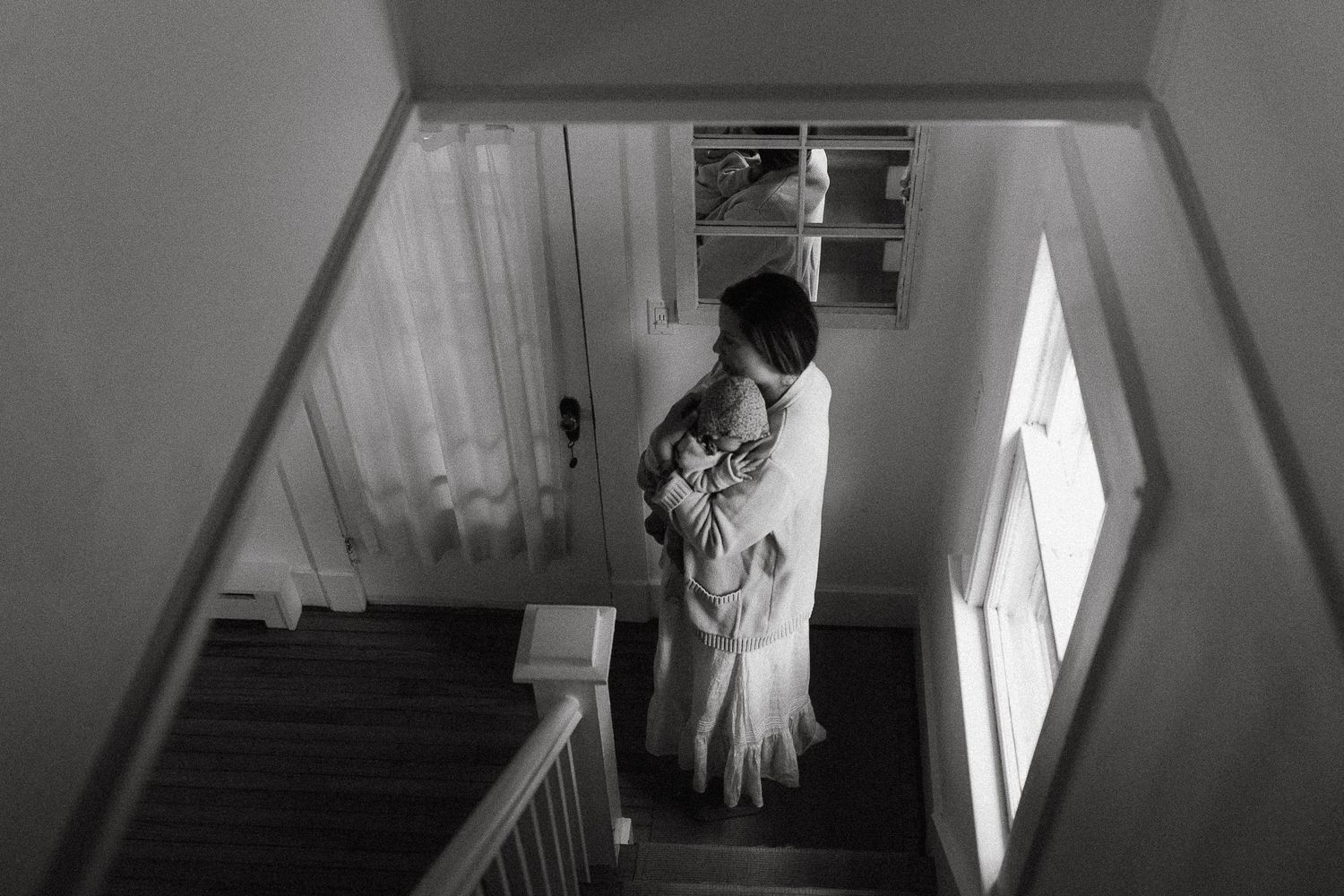 Black and white photo of mother cradling newborn baby on a staircase by a window.