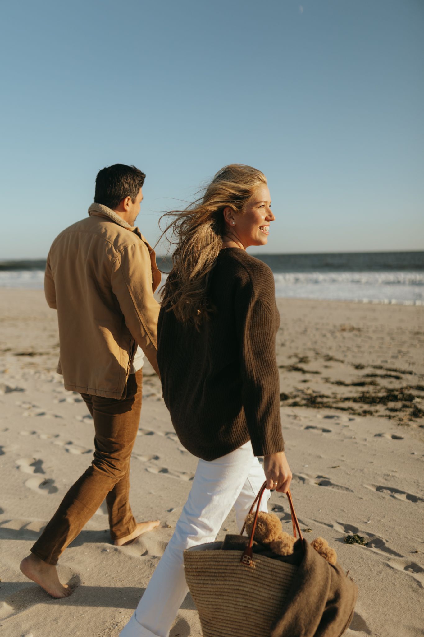 A couple walks along a sunny beach with a straw tote bag.