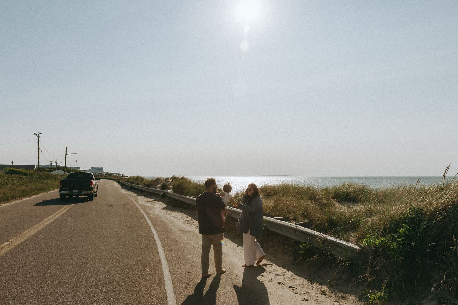 Couple holding a baby standing on a coastal road with ocean views on a sunny day.