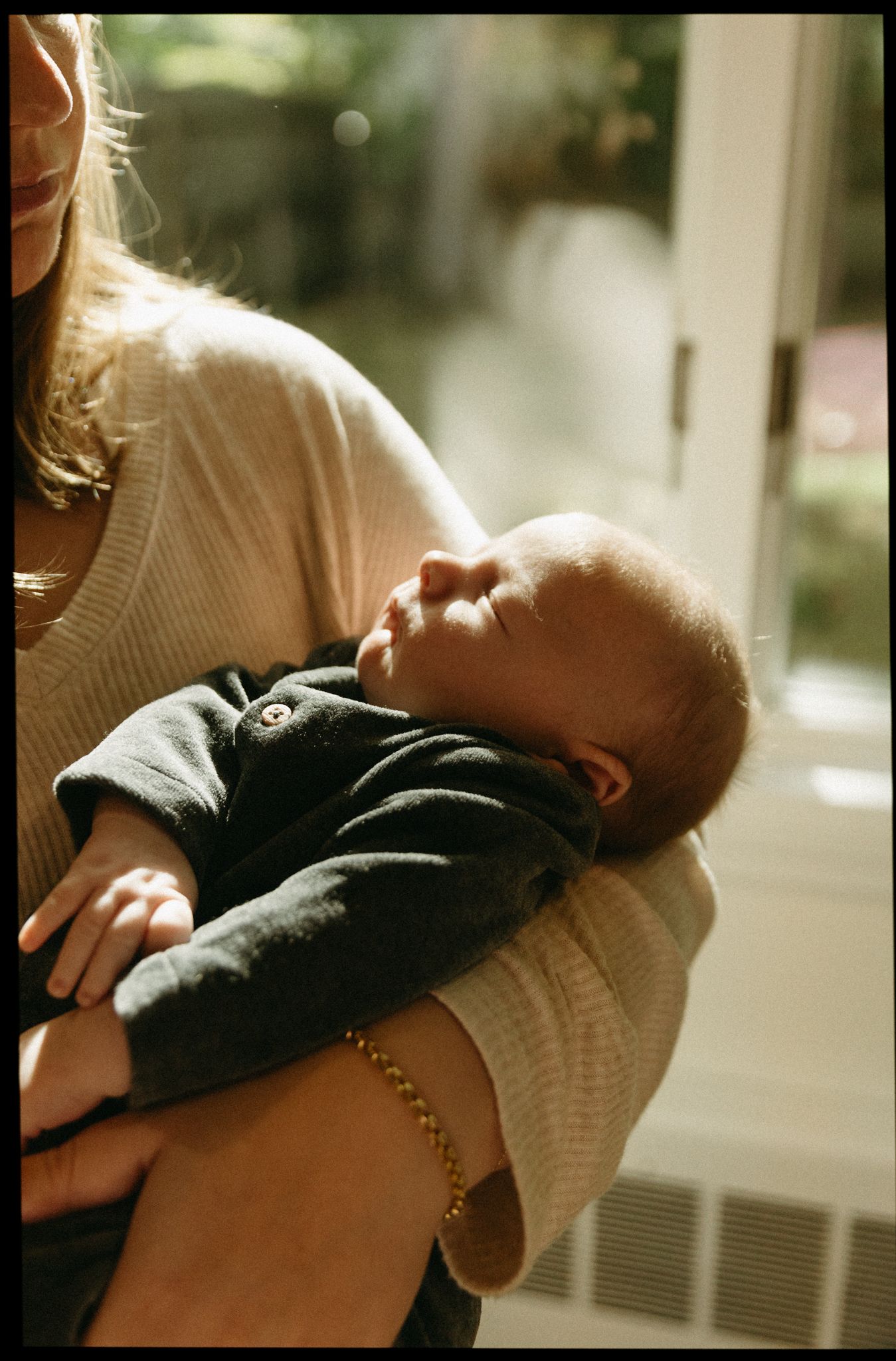 A mother cradling her newborn baby in her arms near a sunlit window, wearing a beige sweater and gold bracelet.