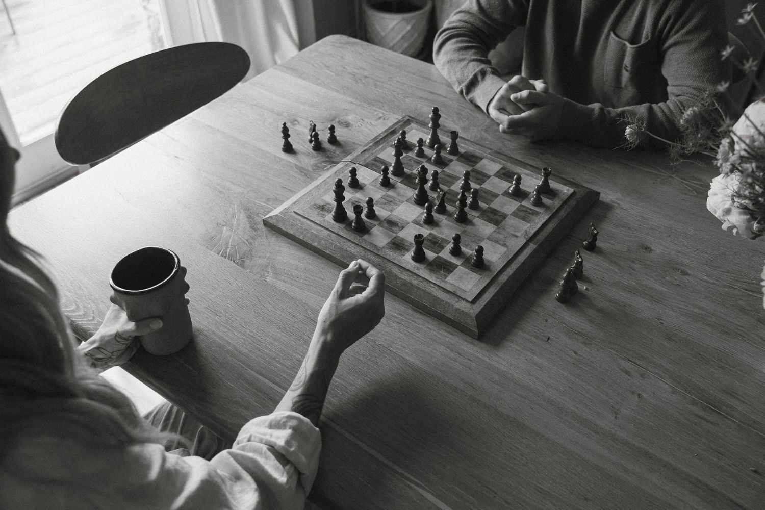 Two people playing chess at a wooden table in black and white photo, one holding a coffee cup.