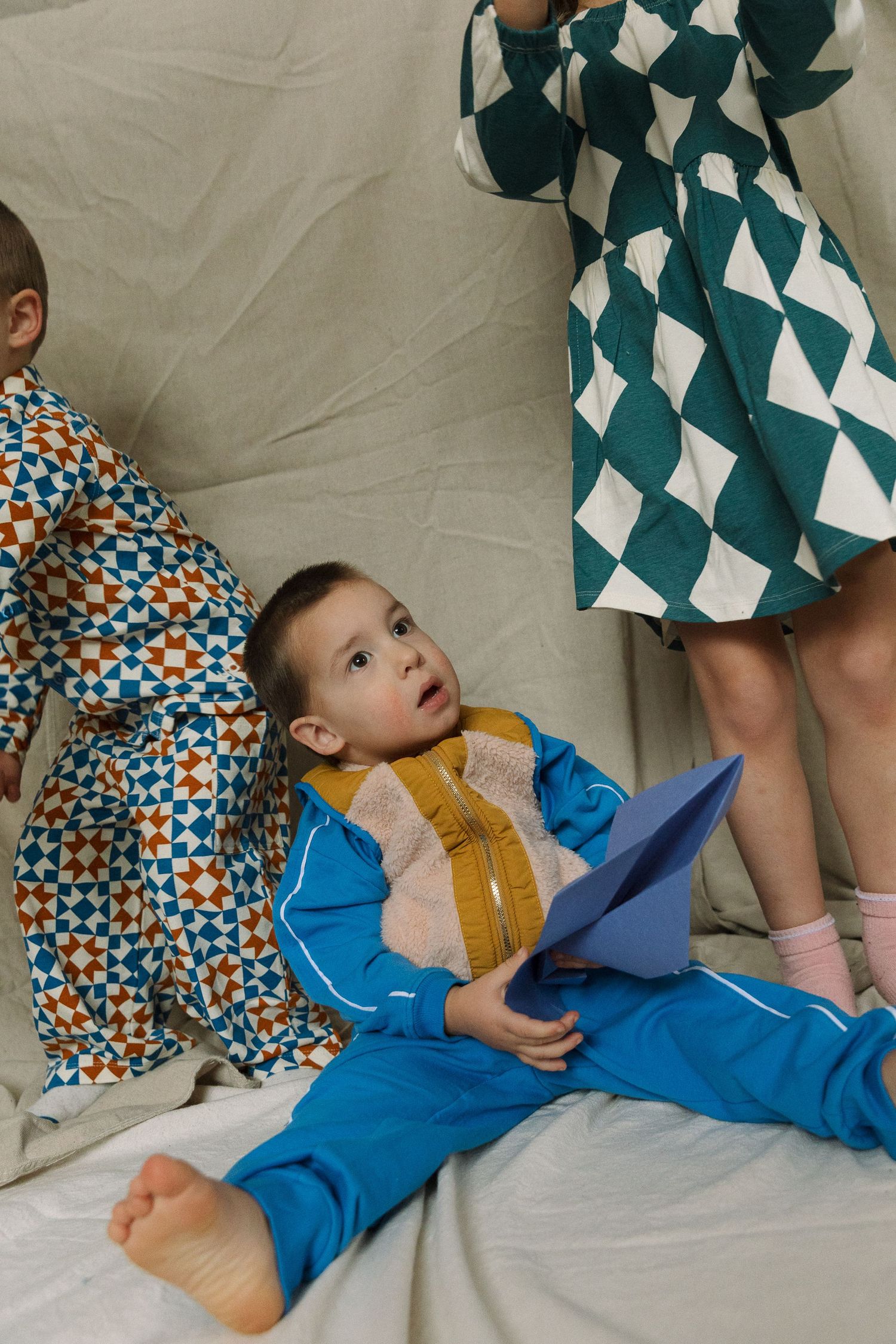Children playing on a beige sofa, wearing colorful patterned outfits, one boy in blue holds a paper airplane.