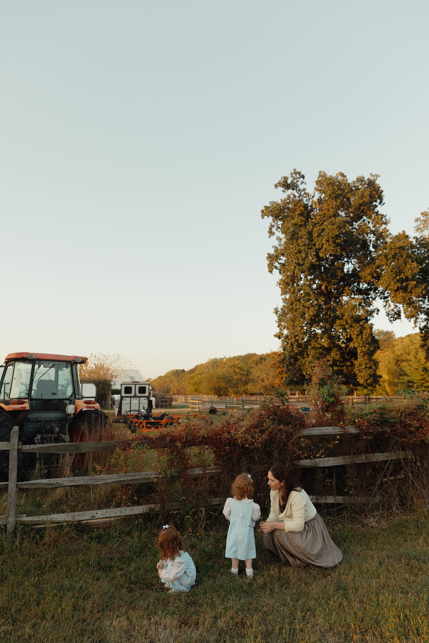 A mother crouches with two young children by a wooden fence on a farm, with a red tractor and autumn trees in the background.