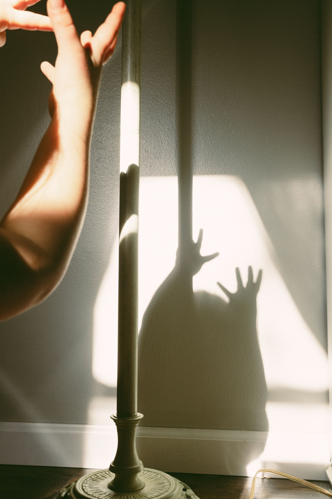 A hand near a lamp casting an eerie shadow with outstretched fingers on a sunlit white wall.