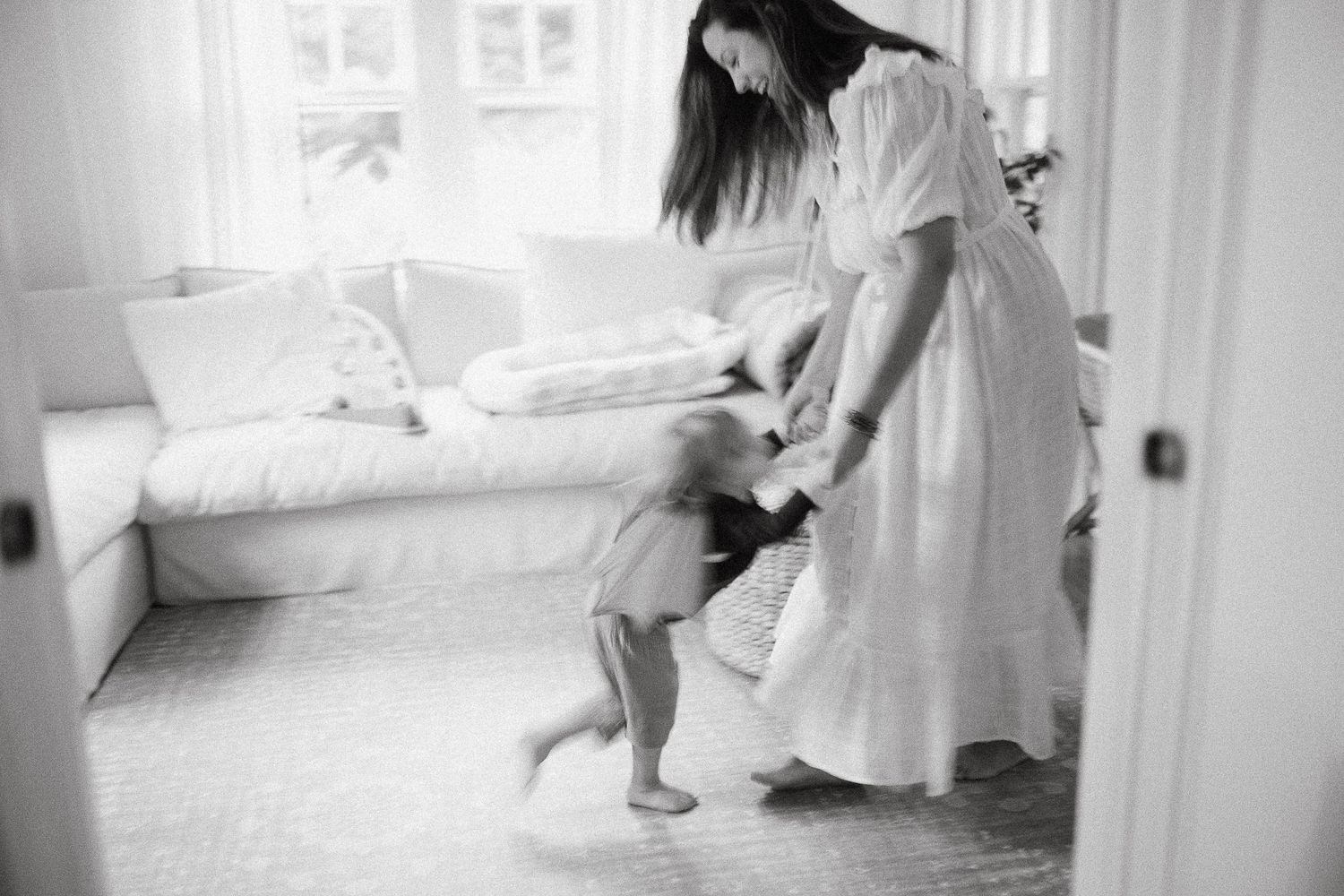 Black and white photo of a mother in white dress dancing with her toddler in a bright room.
