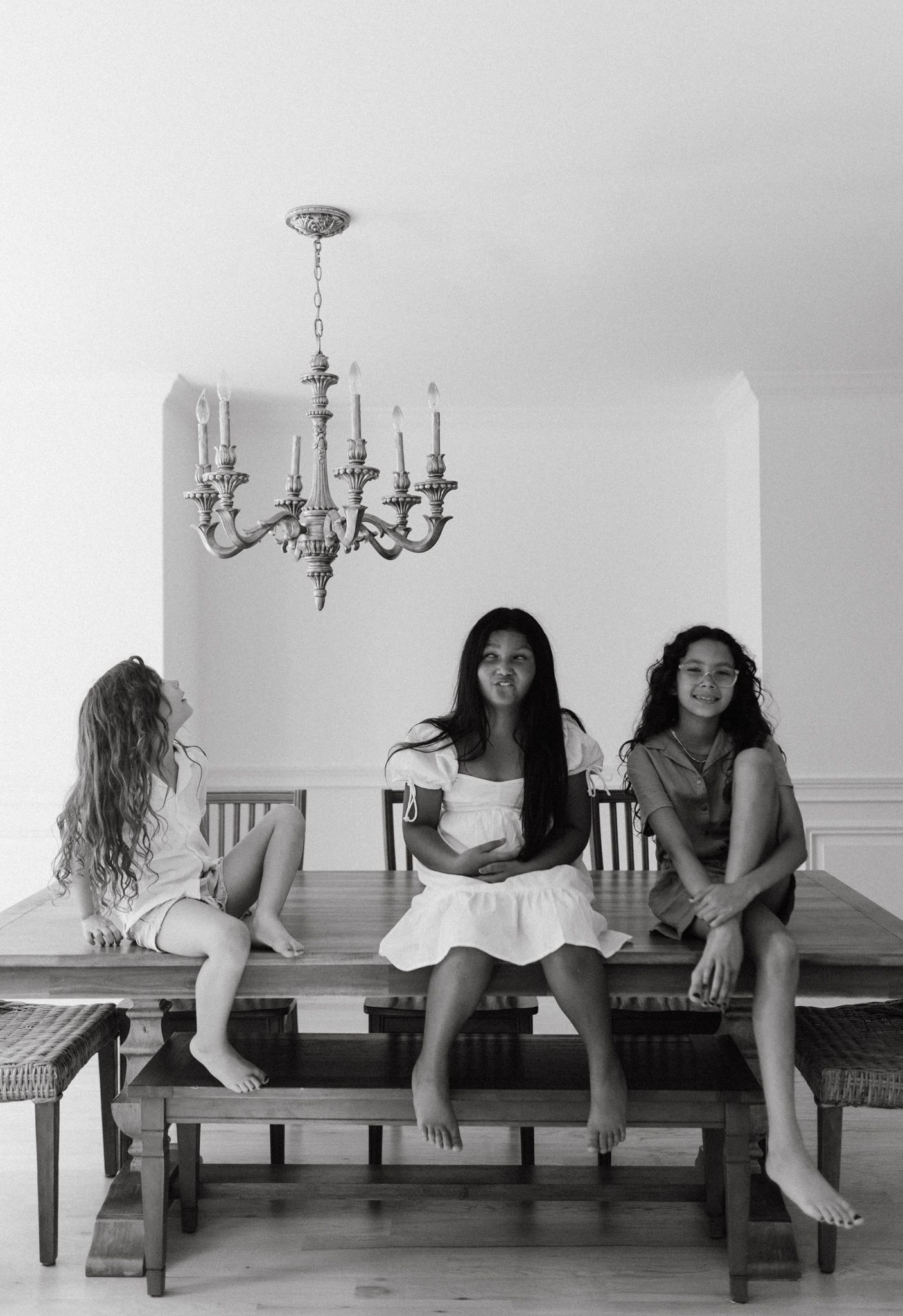 Three friends sitting barefoot on a wooden bench under an elegant chandelier in a bright room.
