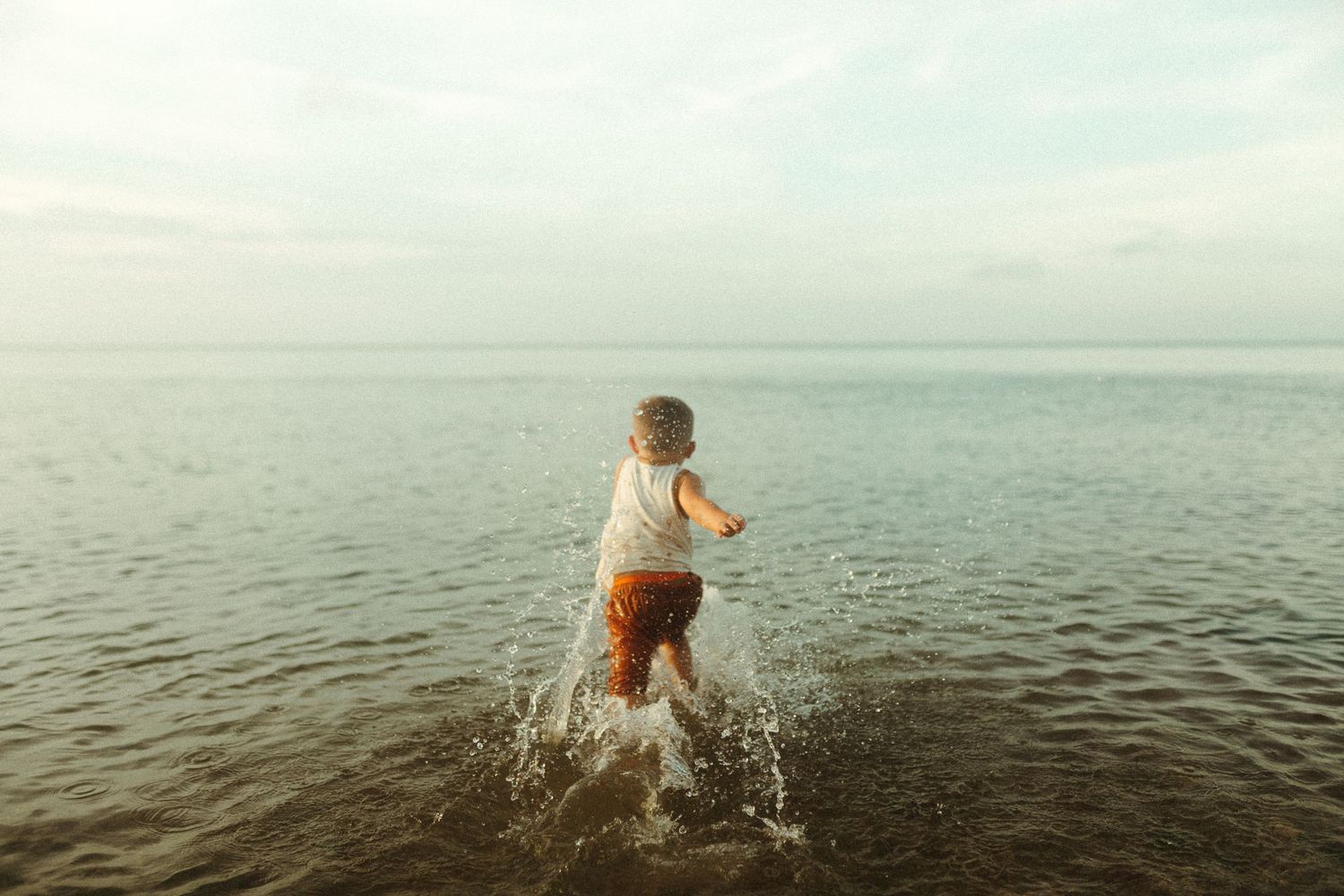 Child splashing in shallow ocean water against cloudy horizon.