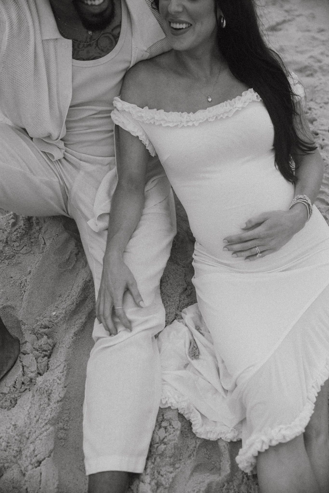 Couple in white clothing sitting casually on sandy beach.