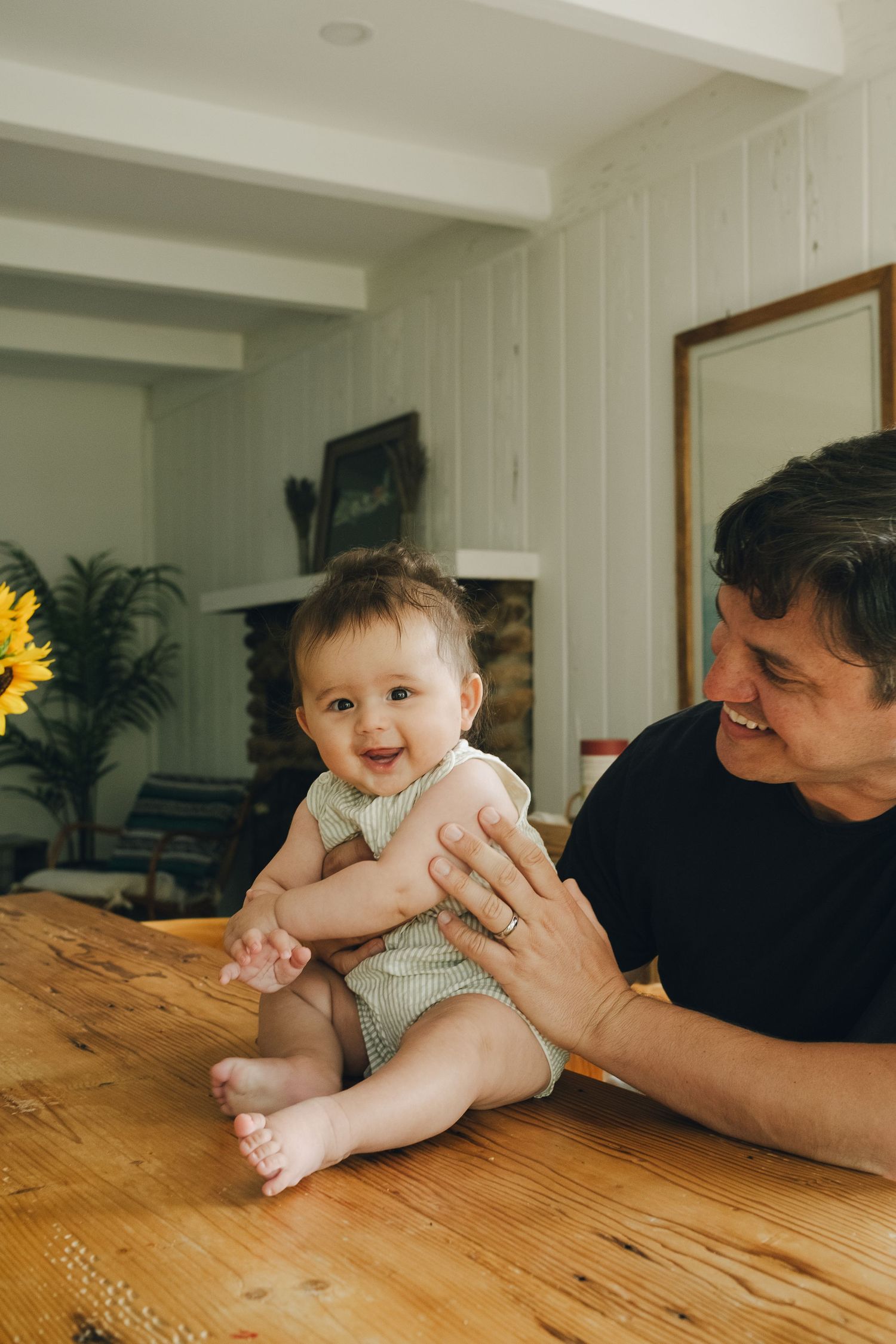 A smiling baby sits on a wooden table while a man in a black shirt plays with them indoors.