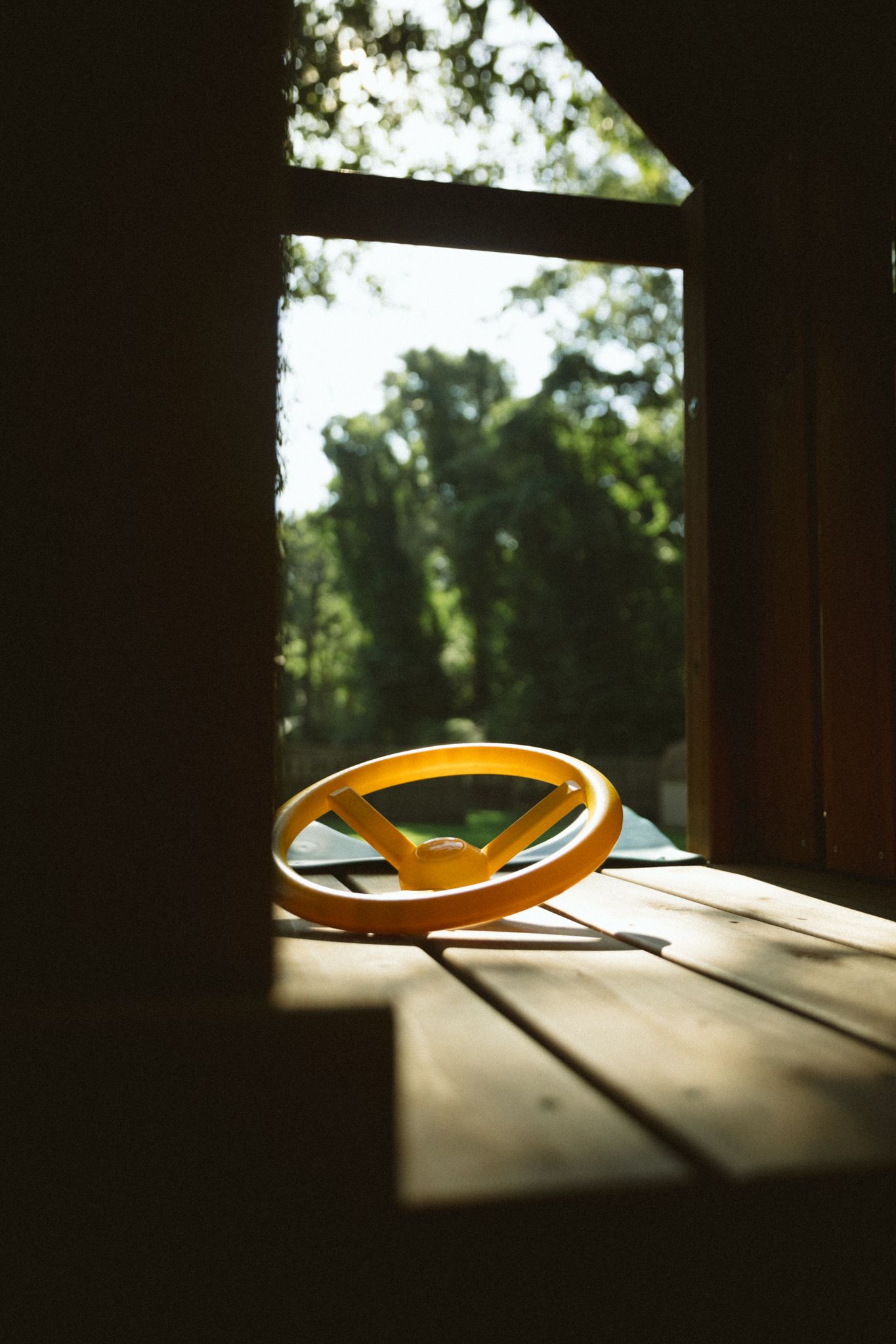 Yellow plastic steering wheel toy resting on wooden deck inside a rustic cabin with green trees visible through window.