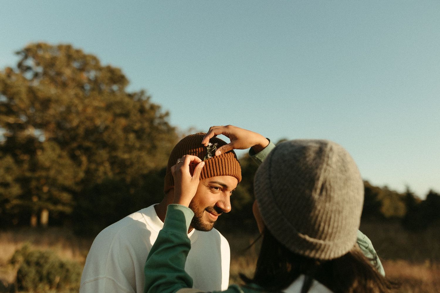 Smiling man wearing a brown beanie hat outdoors in a sunny autumn field with trees behind.
