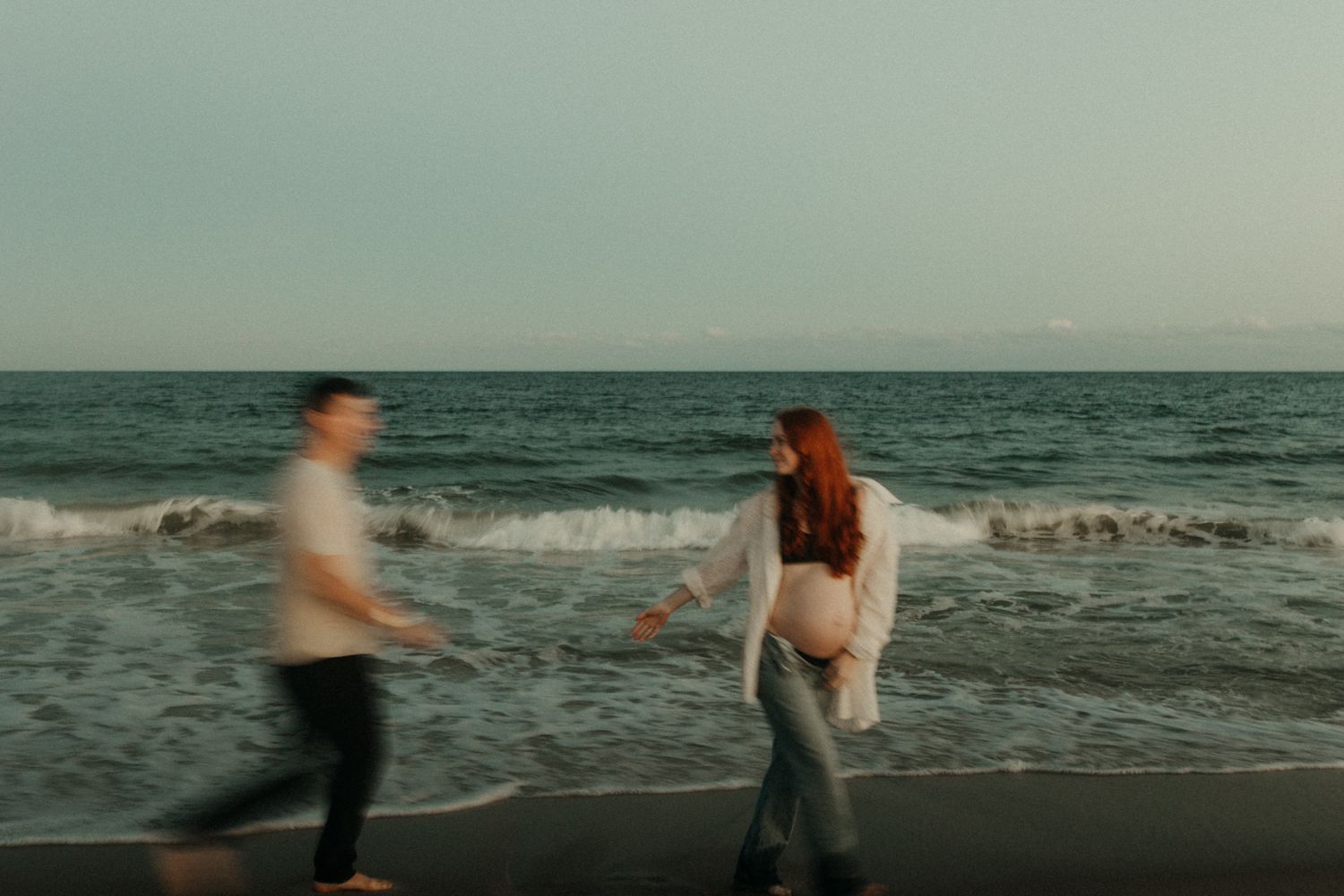 A couple walks hand in hand along a beach shoreline at sunset with crashing waves in the background.