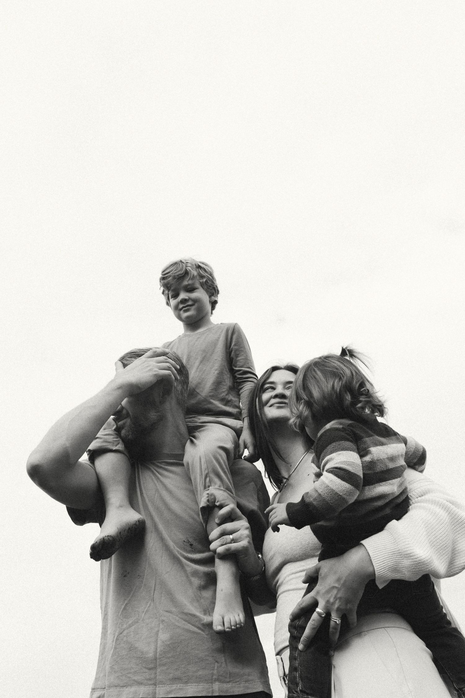 Black and white photo of a family with two children on shoulders, smiling outdoors on a cloudy day.
