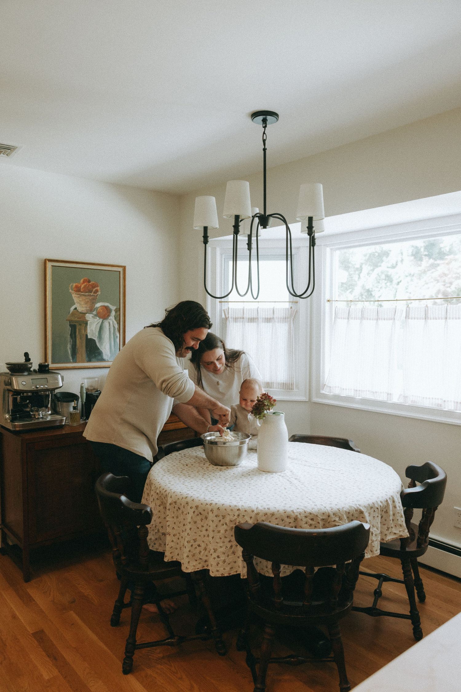 Parent and child preparing food together at round dining table with floral tablecloth indoors.