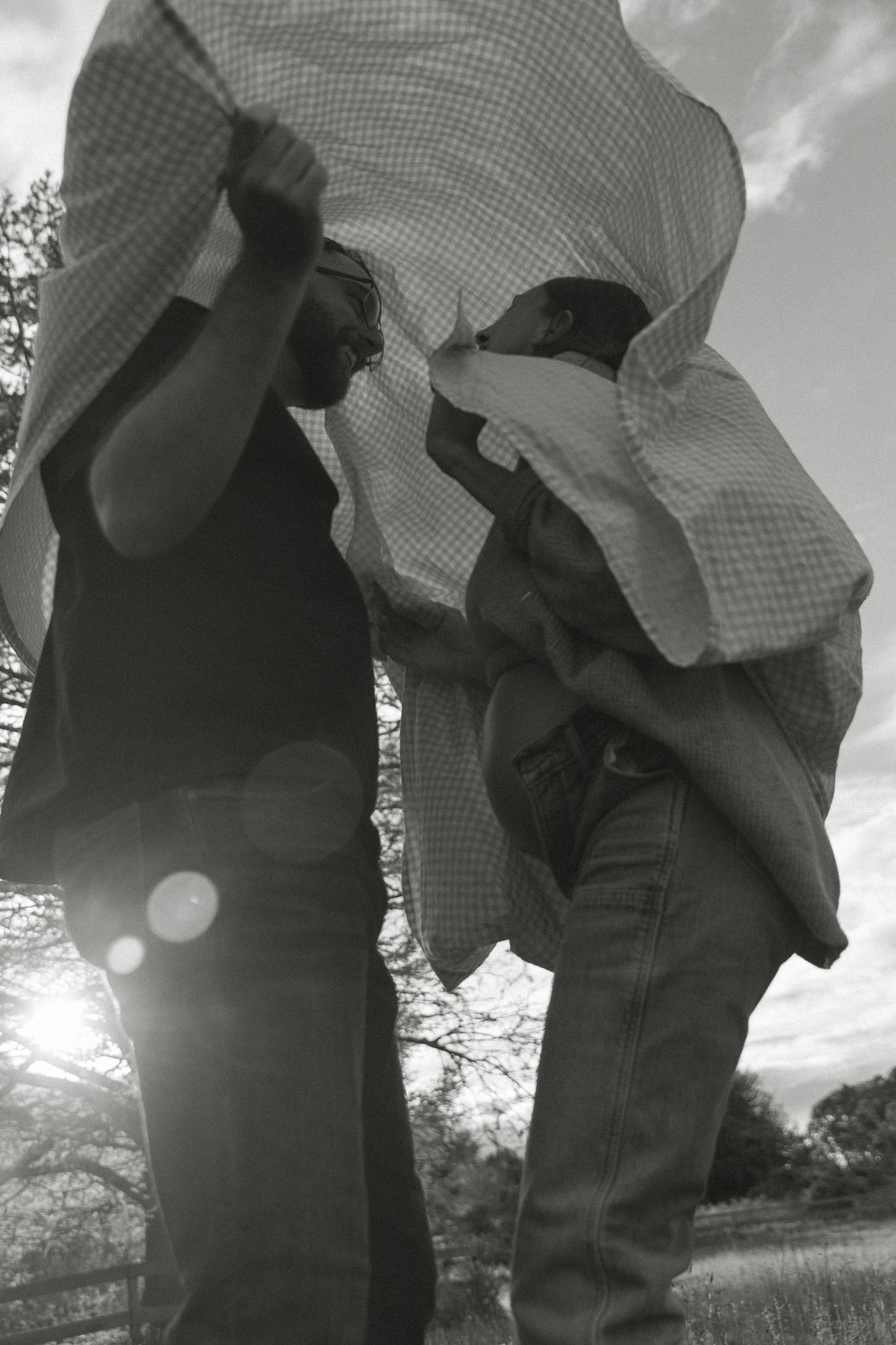 Black and white photo of couple playfully shaking out a blanket outdoors in bright sunlight.
