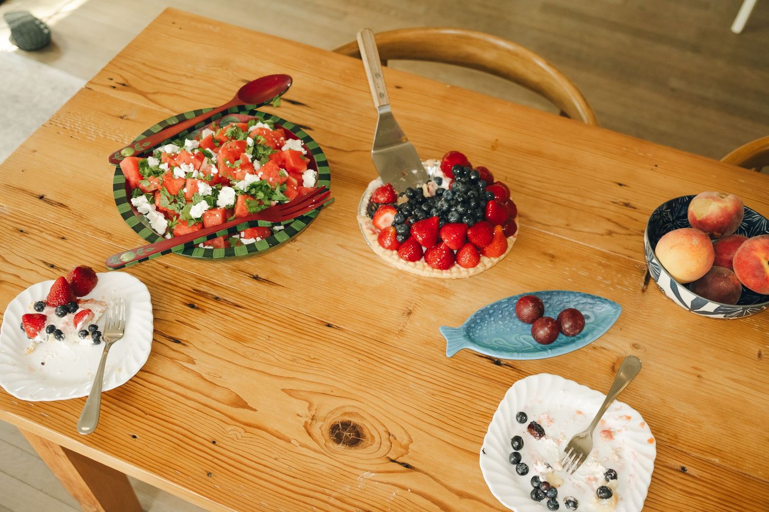 Wooden dining table with fresh fruit tart, watermelon feta salad, berry plates, and peaches in a bowl.