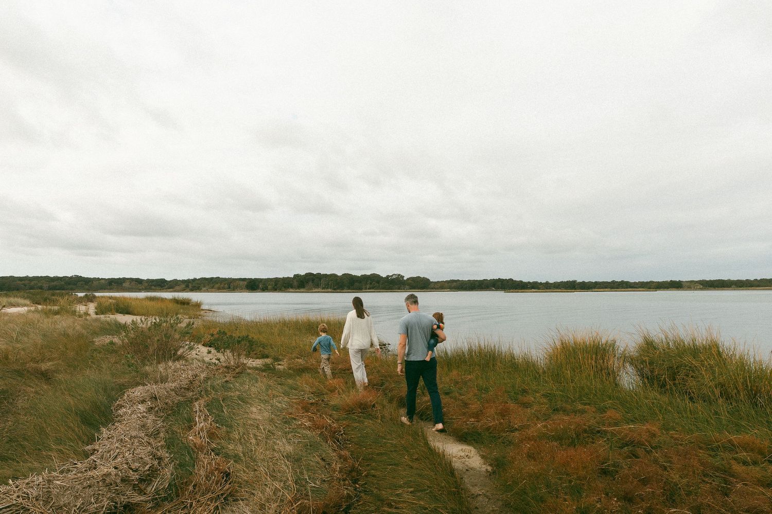 Family walking along a scenic coastal path with tall grasses and a calm lake under an overcast sky.