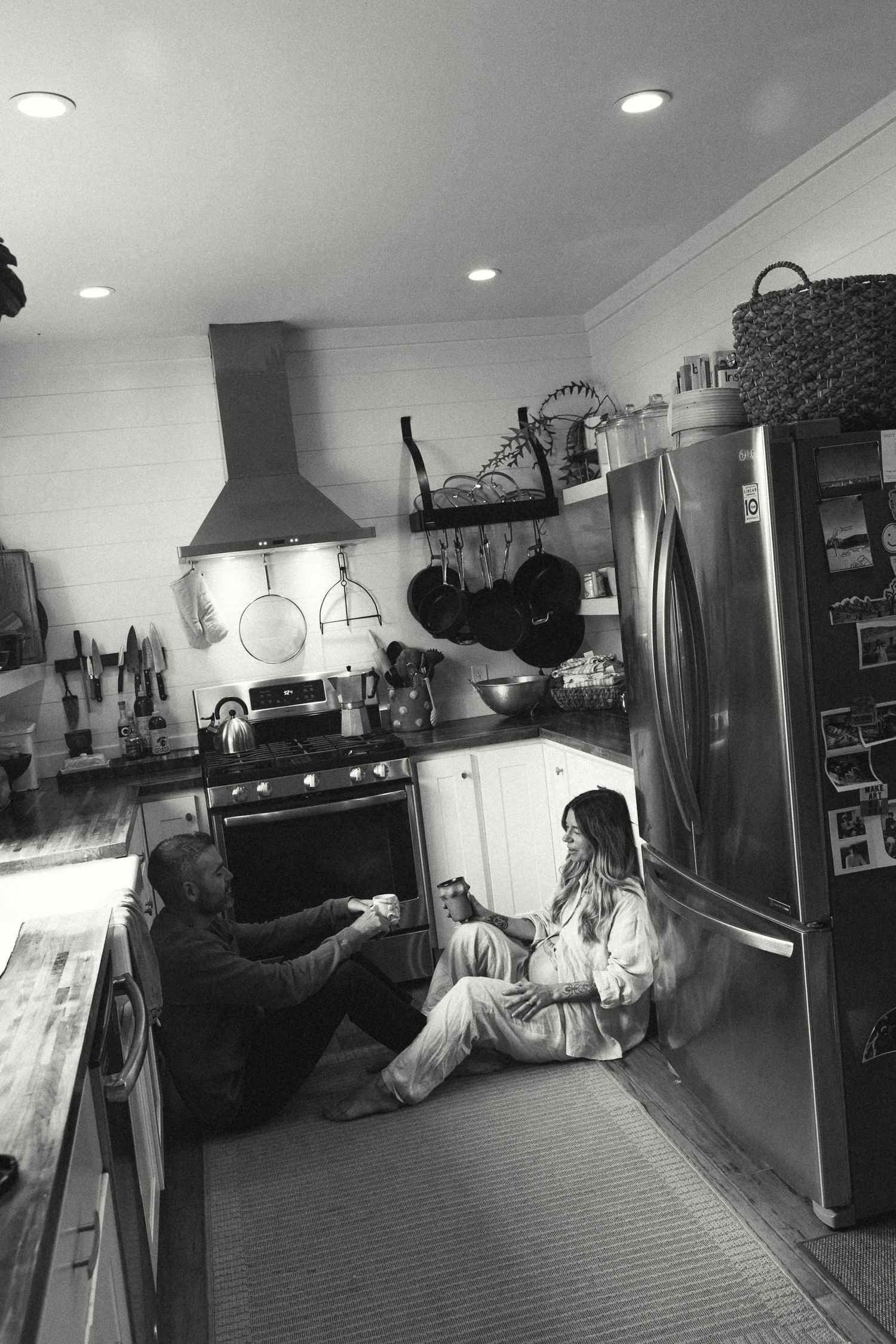 Couple sitting on kitchen floor sharing drinks in cozy black and white home photo.