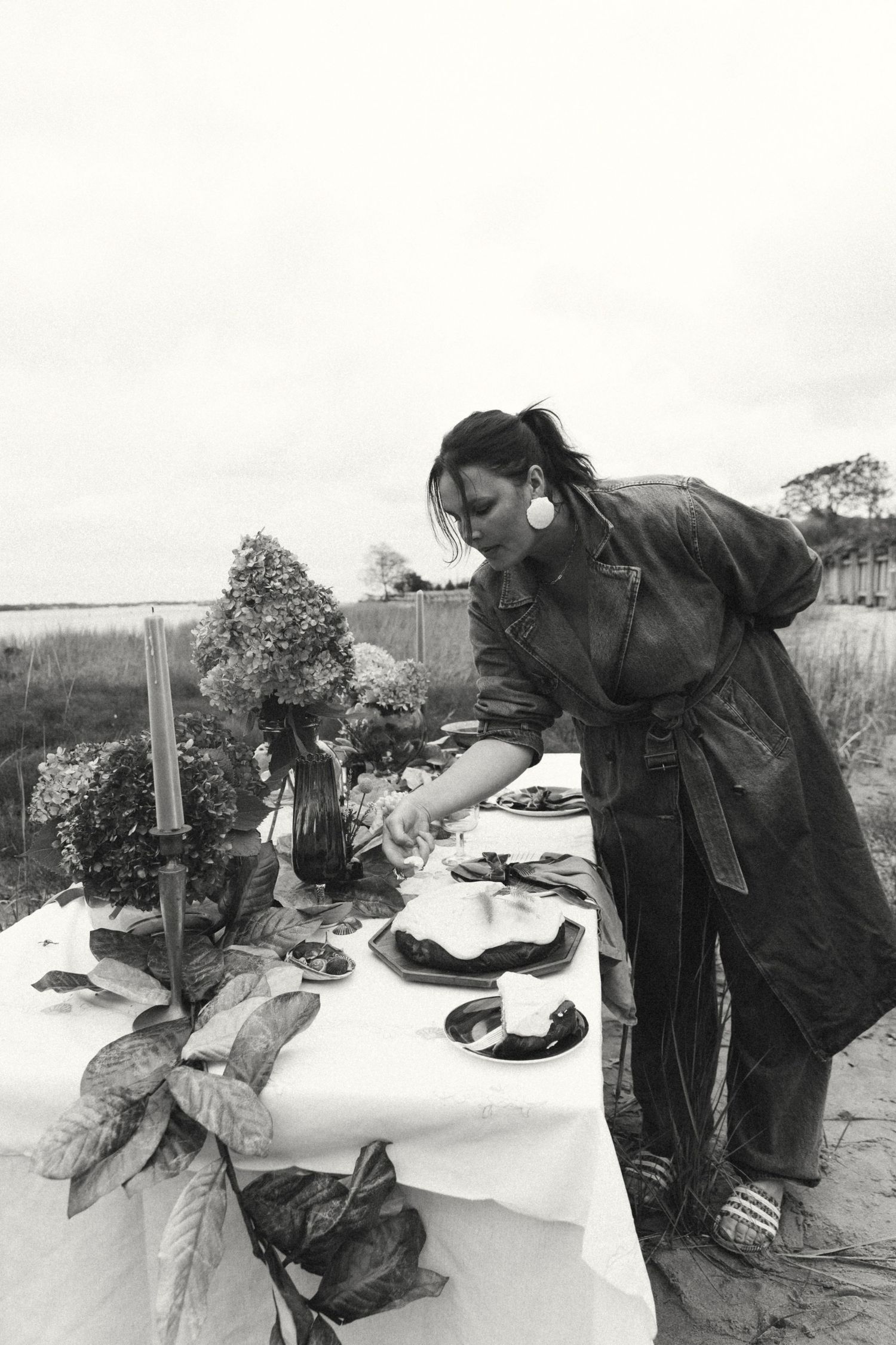 Woman in overalls arranging an outdoor tablescape with flowers and candles in a black and white photo.
