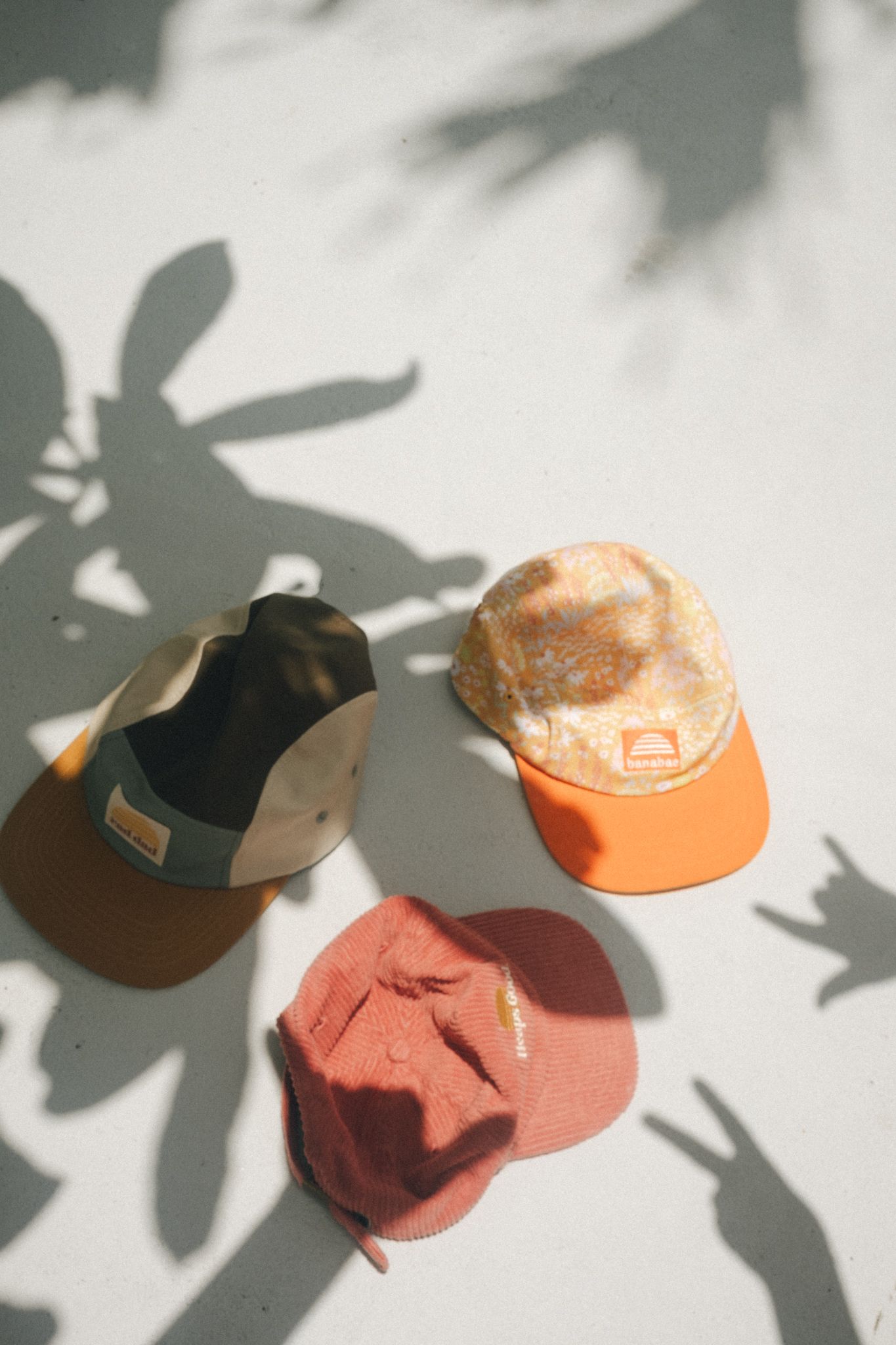 Three colorful caps in olive, orange, and red arranged on a light surface with leafy shadows.