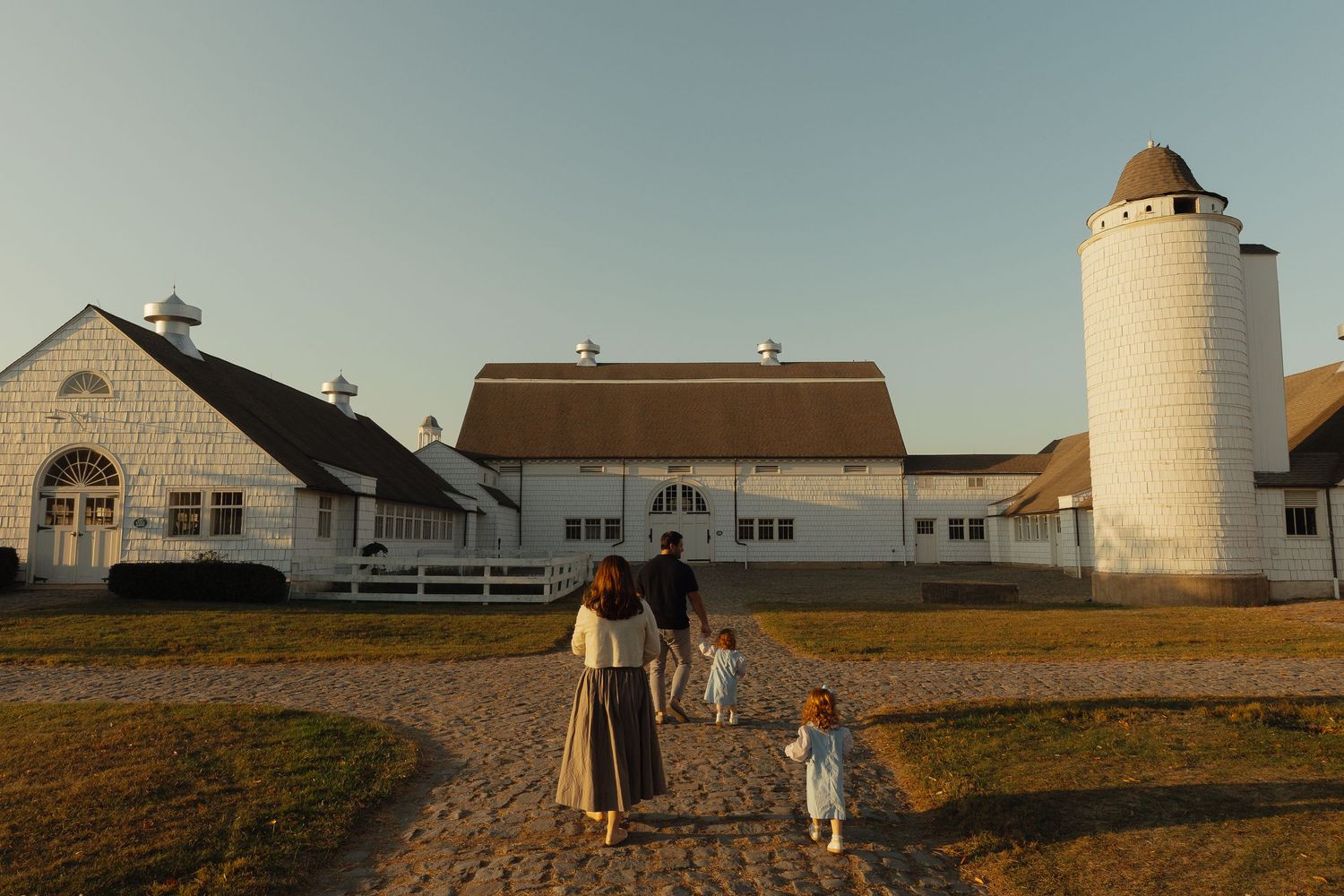 Family walking toward a white farmhouse with silo at golden hour on a sandy path.