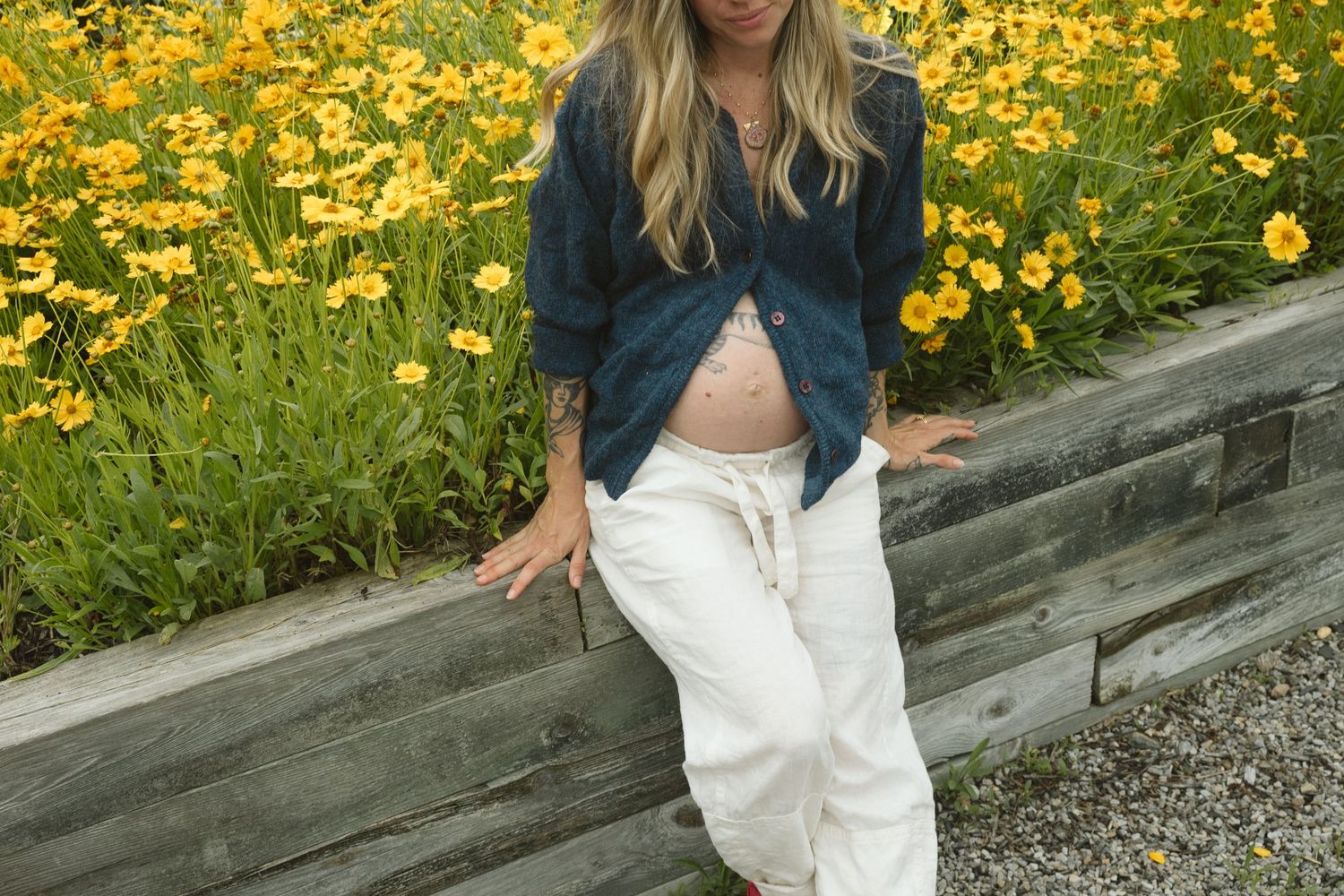 Pregnant woman in denim shirt and white pants poses outdoors near yellow wildflowers and wooden border.
