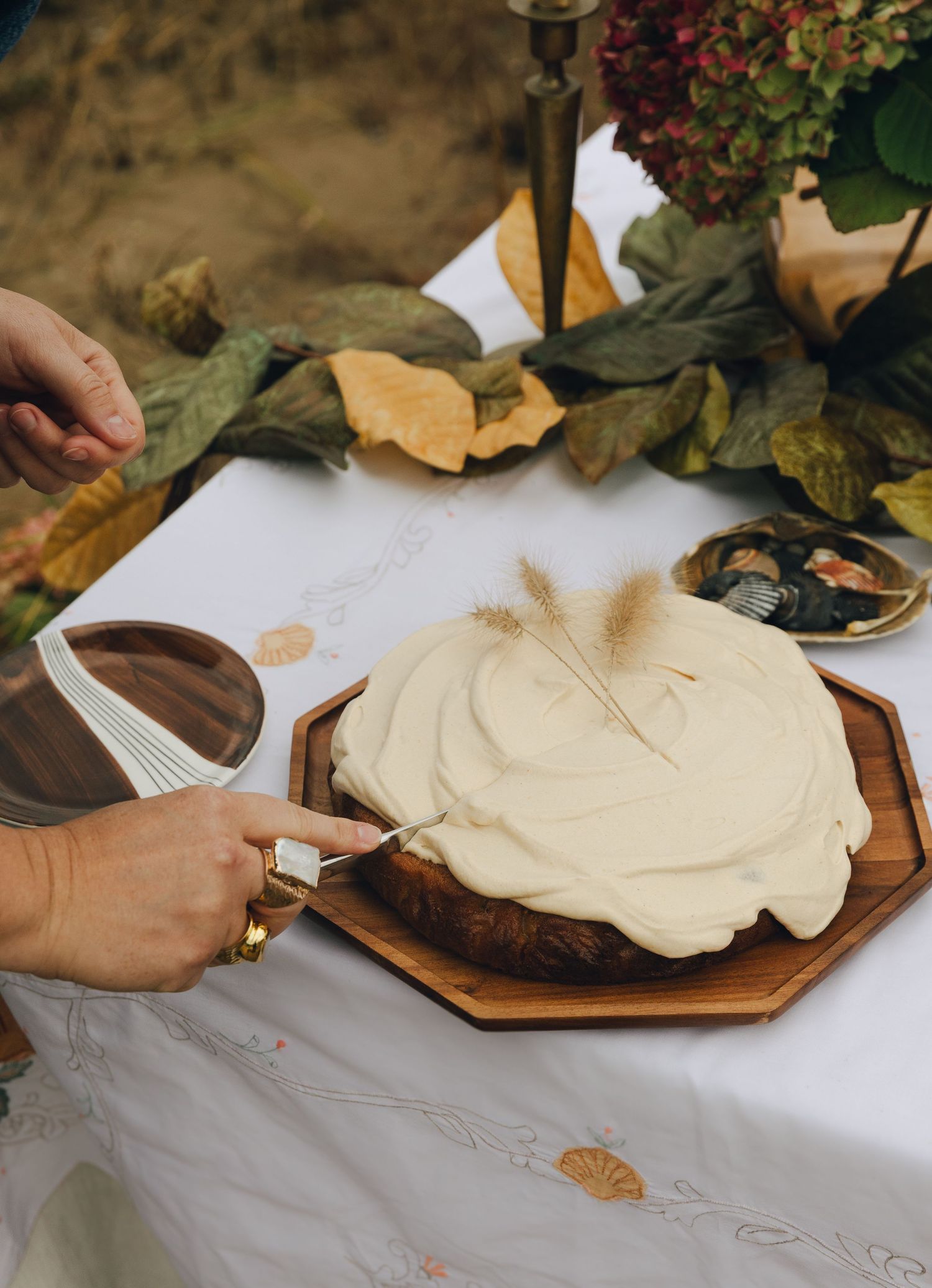 Hands cutting a cream frosted naked cake on wooden board with autumn leaves and floral decor on white tablecloth.