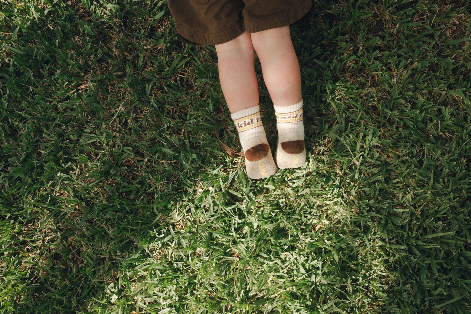Close-up of a child's feet in white socks and brown Mary Jane shoes standing on lush green grass.