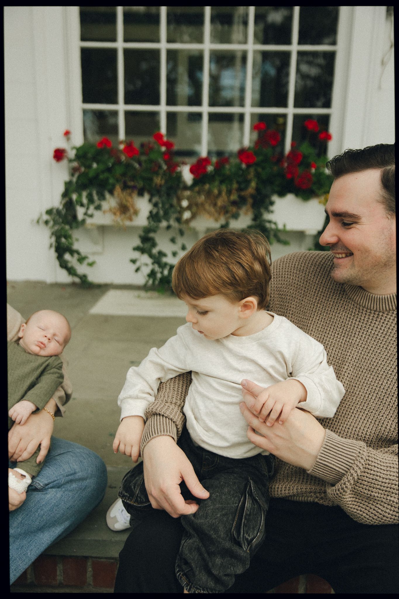 A smiling father holds a toddler on his lap outdoors, with a sleeping newborn nearby and red flowers in background.