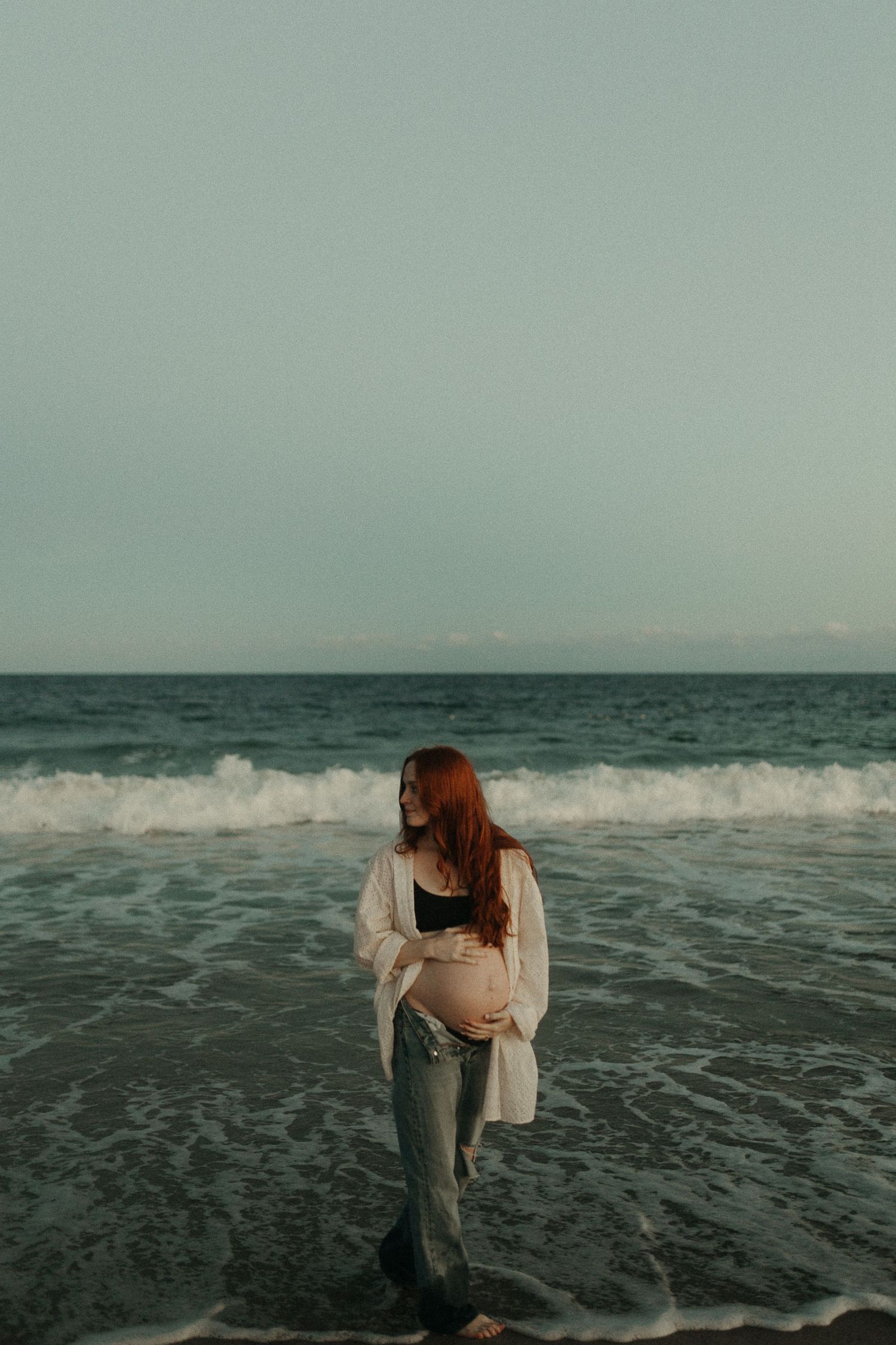 A person in jeans and white top walks along the beach with waves crashing behind.