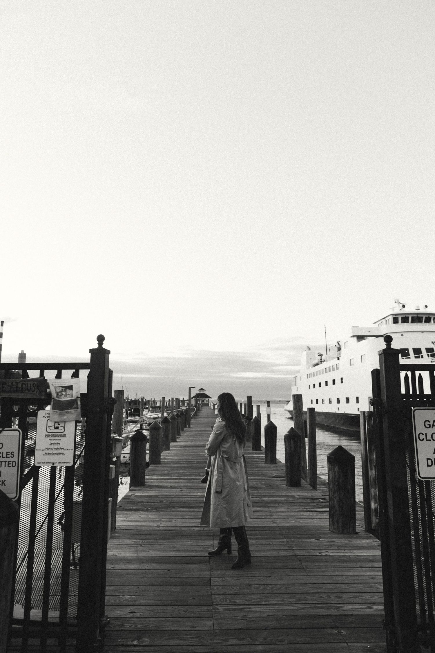 A black and white photo of people walking on a long wooden pier with a ferry in the distance.